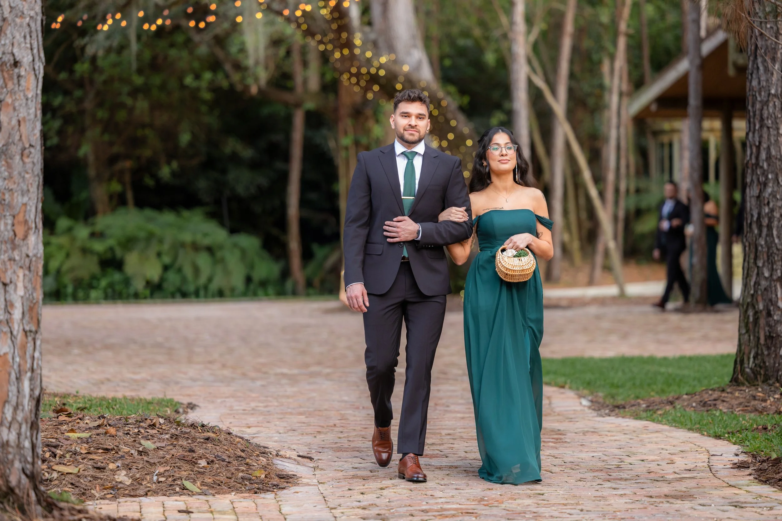 A couple dressed in formal attire walking arm in arm on a brick path in a wooded outdoor setting, with fairy lights hanging in the background.