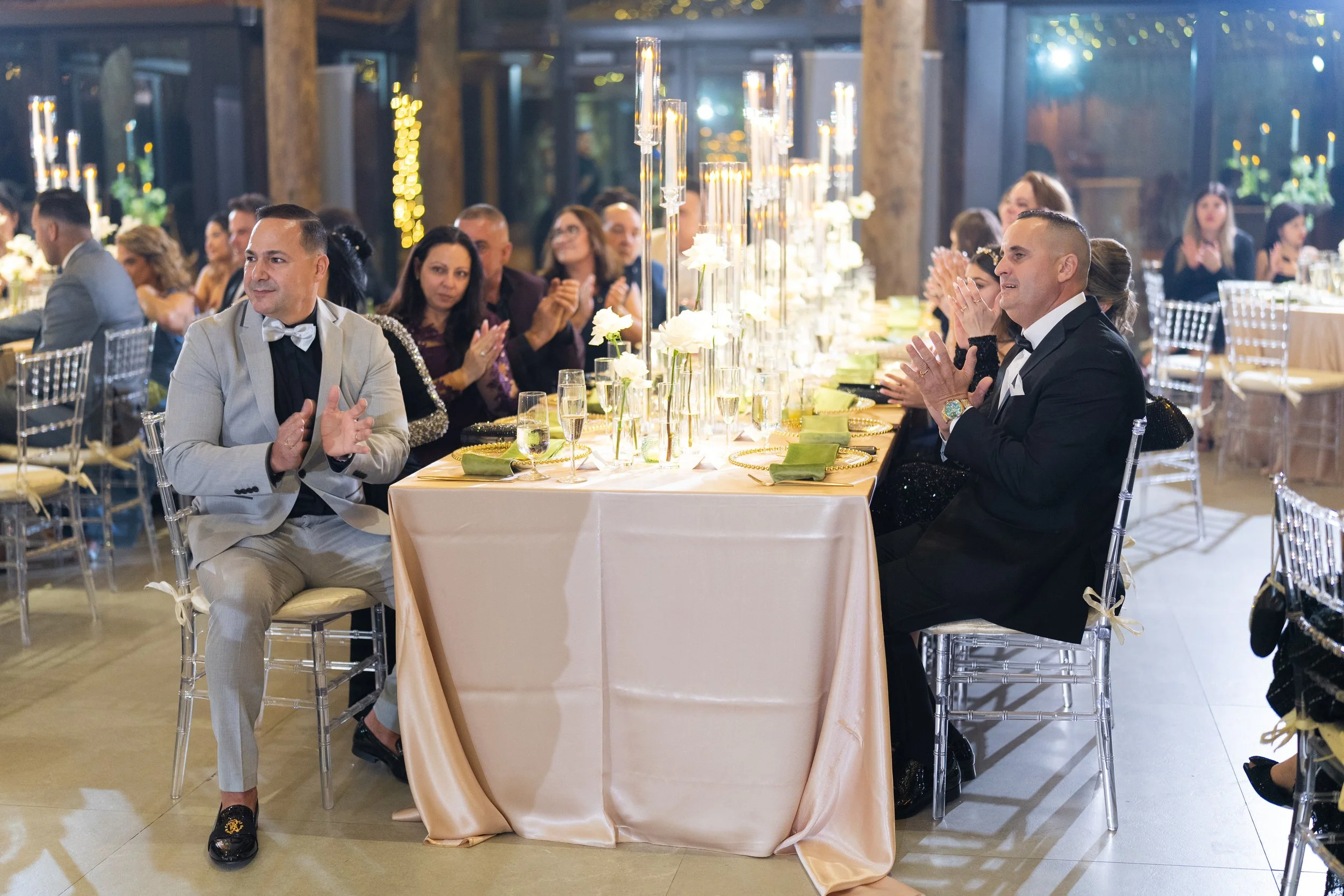 People dressed in formal attire seated at a long banquet table, clapping during a celebration, with decorative flower centerpieces and candle-like lights.