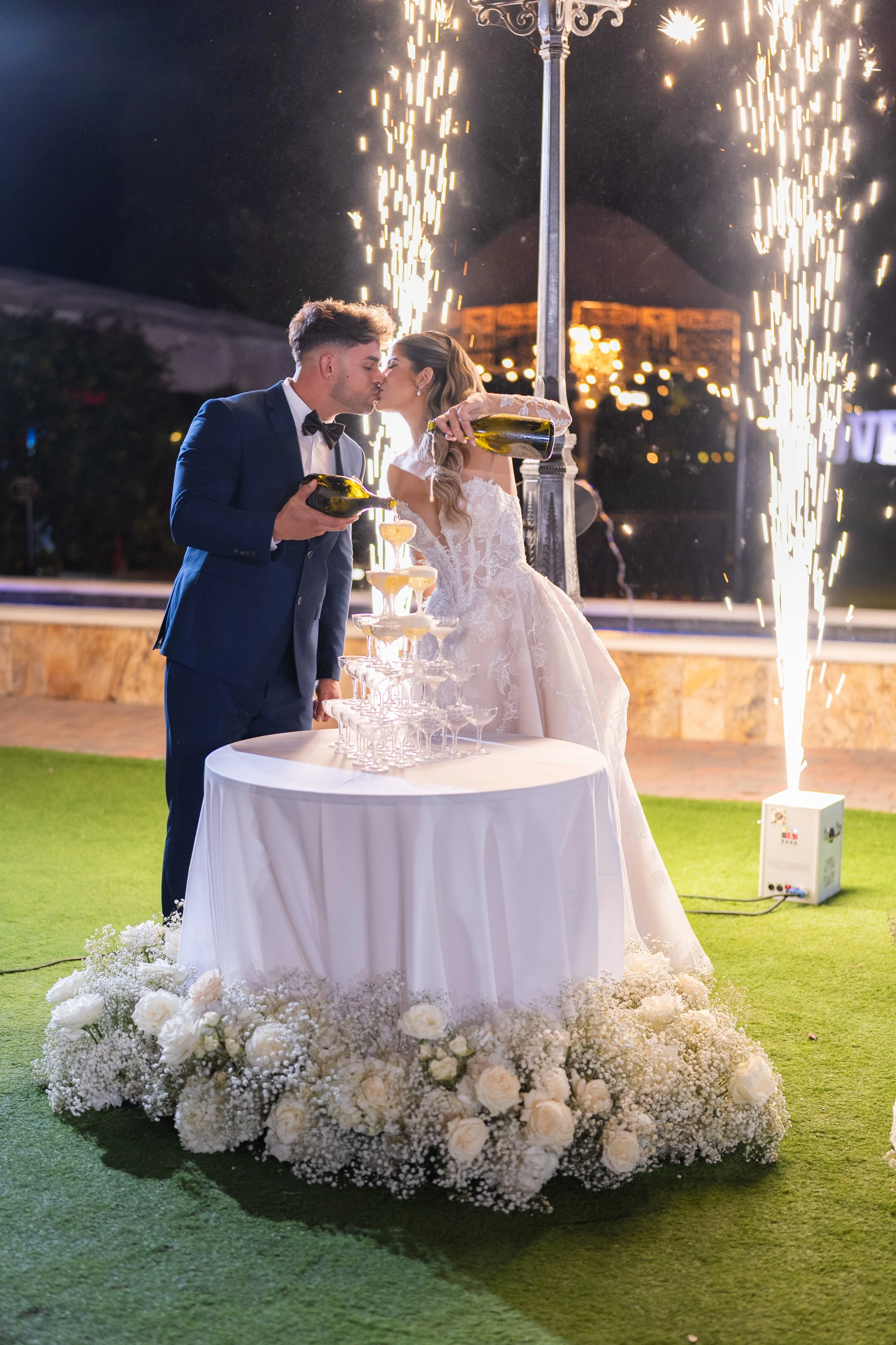 A bride and groom in wedding attire in a nighttime celebration, pouring champagne into a tower of glasses with sparklers and fireworks behind them, surrounded by white floral arrangements.