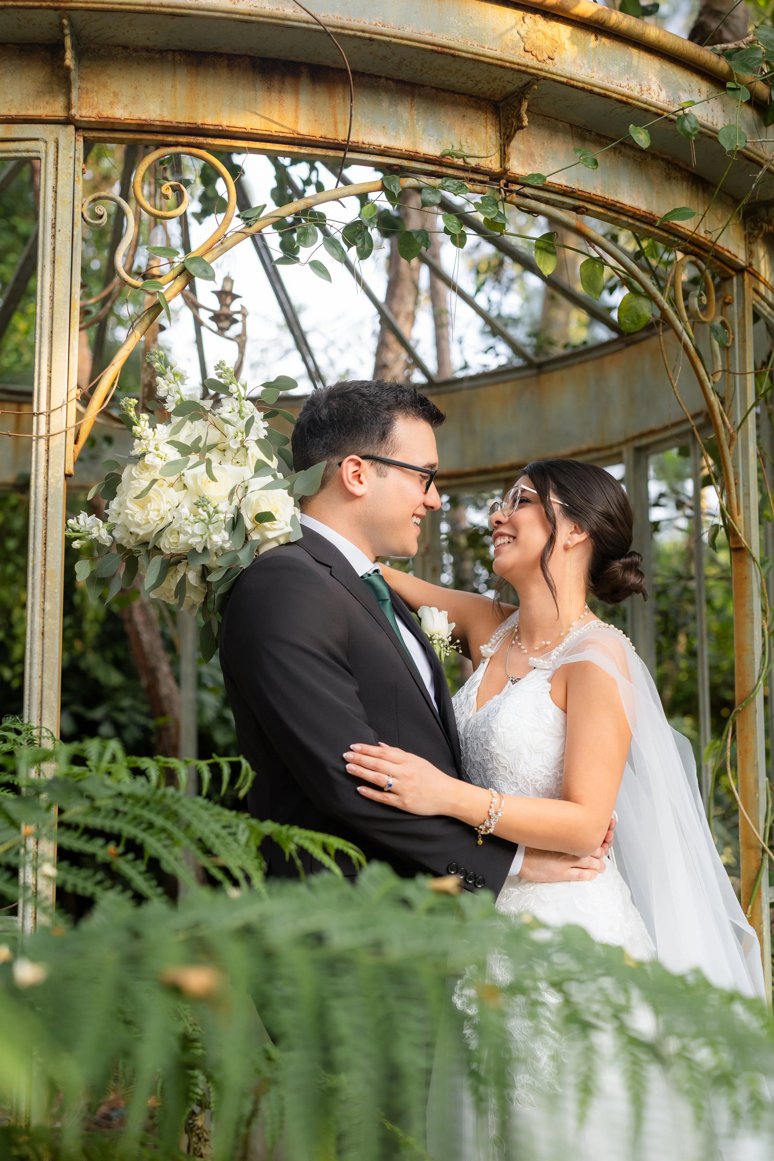 A bride and groom share a romantic moment during their wedding, standing close and looking into each other's eyes beneath a rustic, vine-covered metal arch.