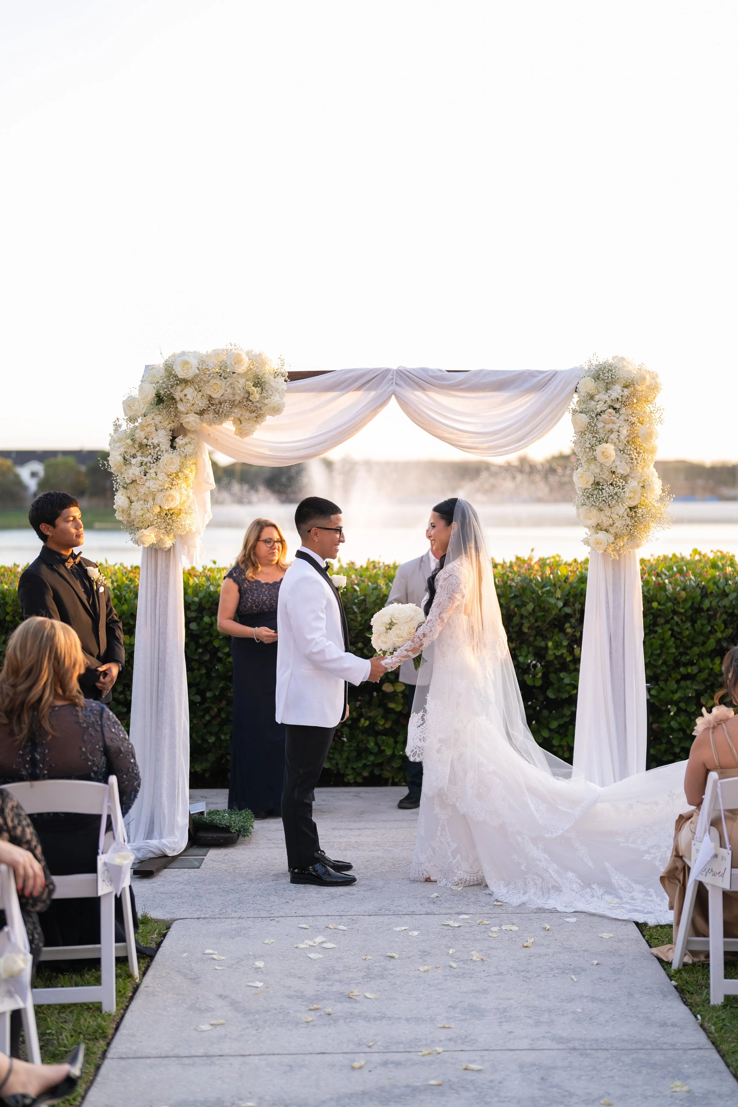 A wedding ceremony taking place outdoors by a lake with a fountain in the background. The bride and groom stand under a floral arch, holding hands. The bride is in a white lace wedding dress and veil, holding a bouquet of white flowers. The groom is 