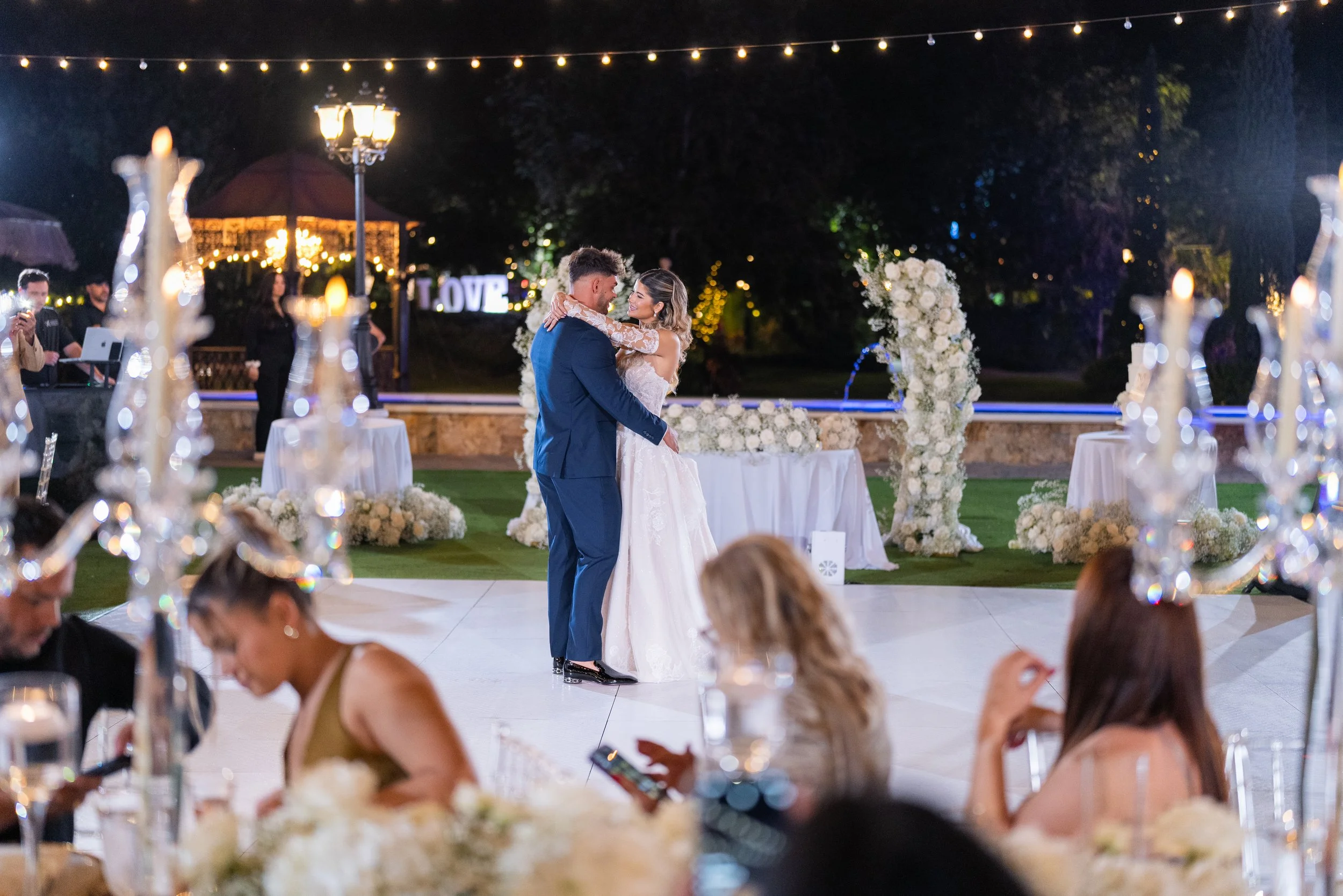 A bride and groom share a dance at their wedding reception outdoors at night, with string lights overhead and guests seated at tables decorated with white flowers.