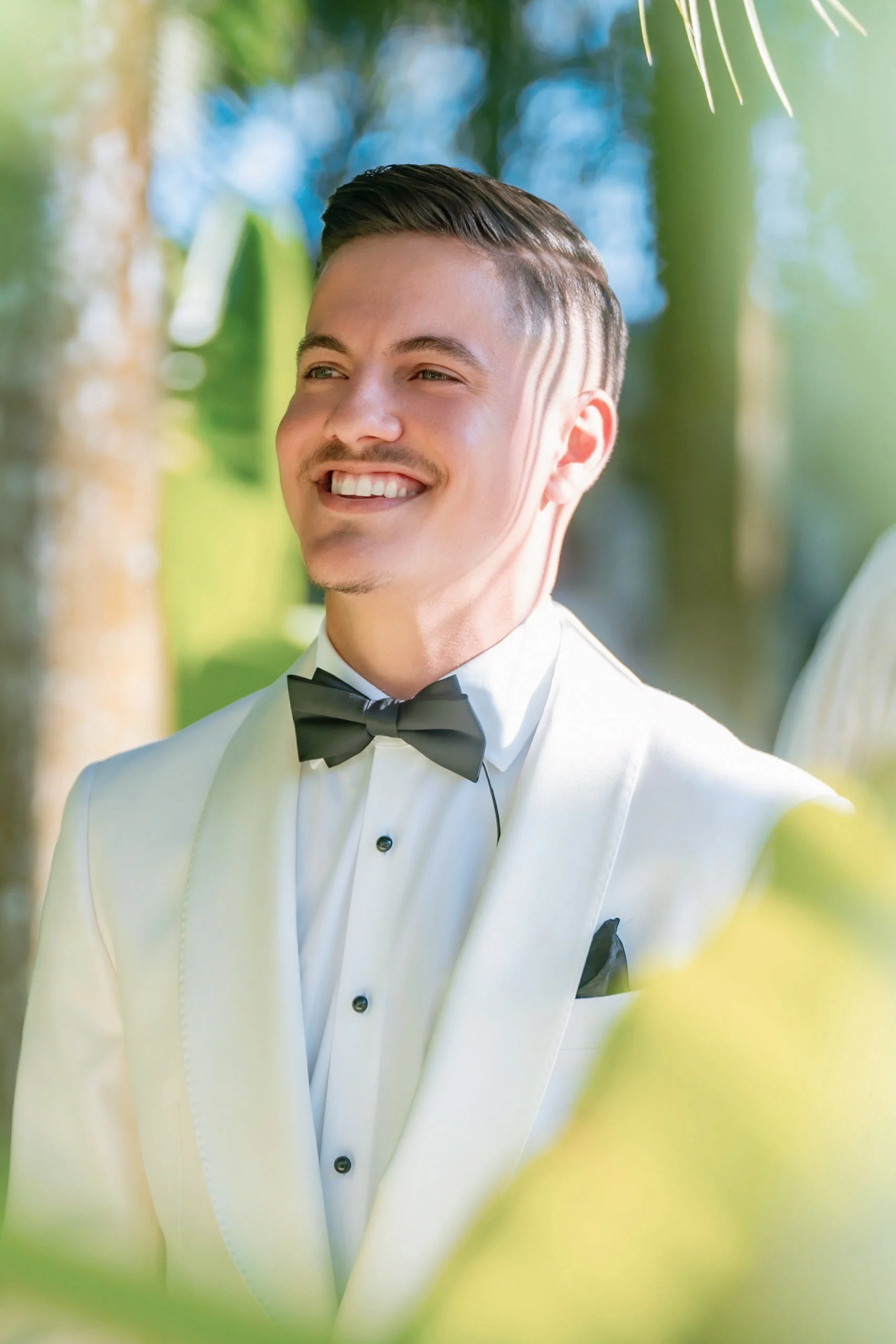 A smiling man wearing a tuxedo with a bow tie outdoors with greenery and blue sky in the background.