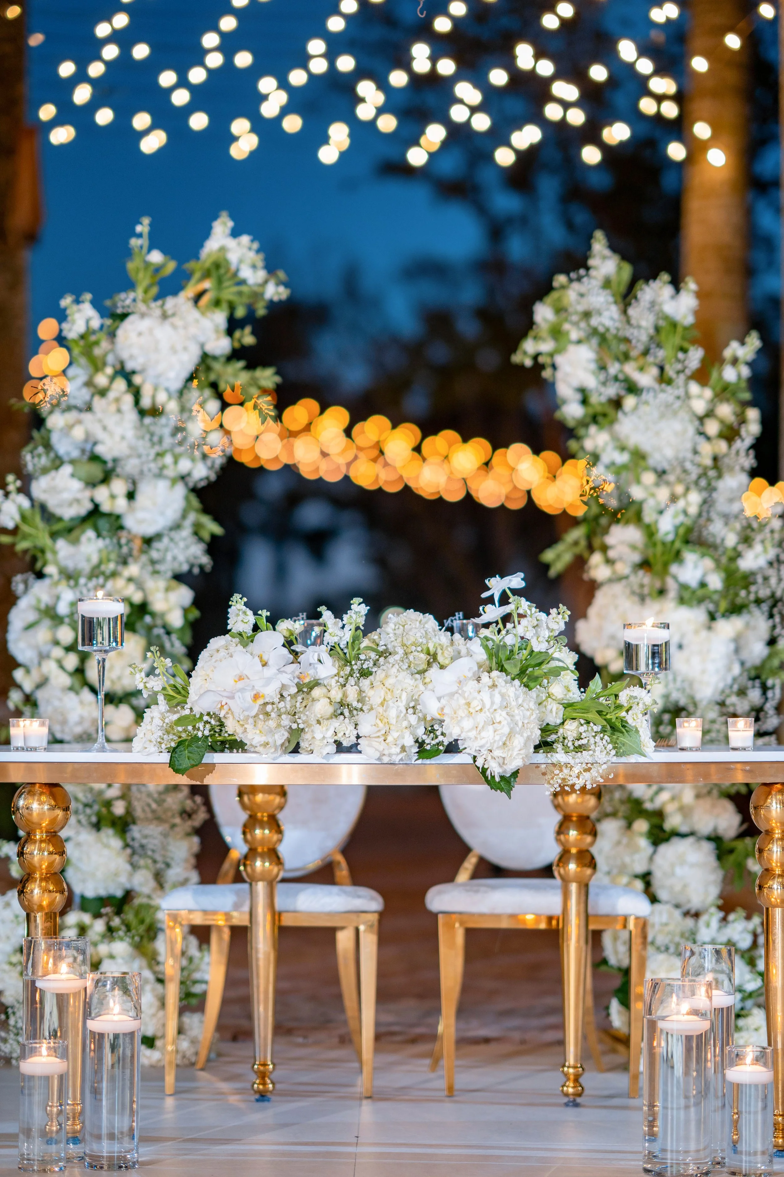 Elegant wedding or event table setup decorated with white flowers and candles, with string lights hanging in the background during dusk or evening.