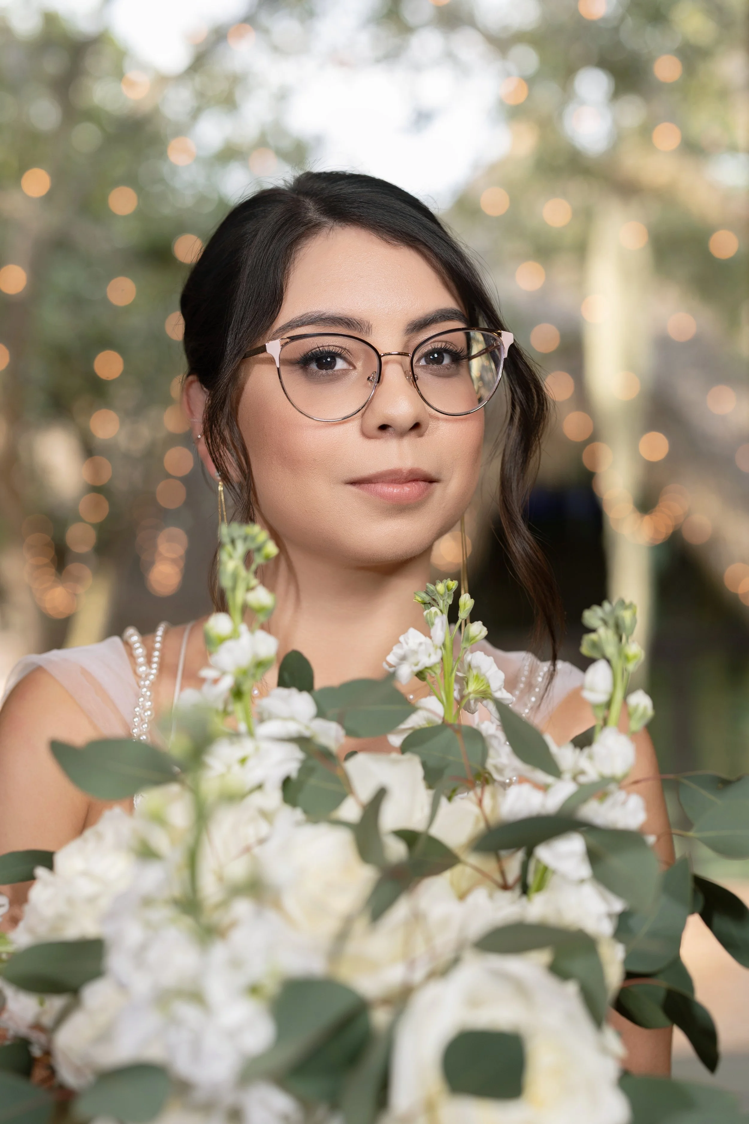Portrait of a woman with glasses looking at camera, holding white flowers, with warm outdoor bokeh background.