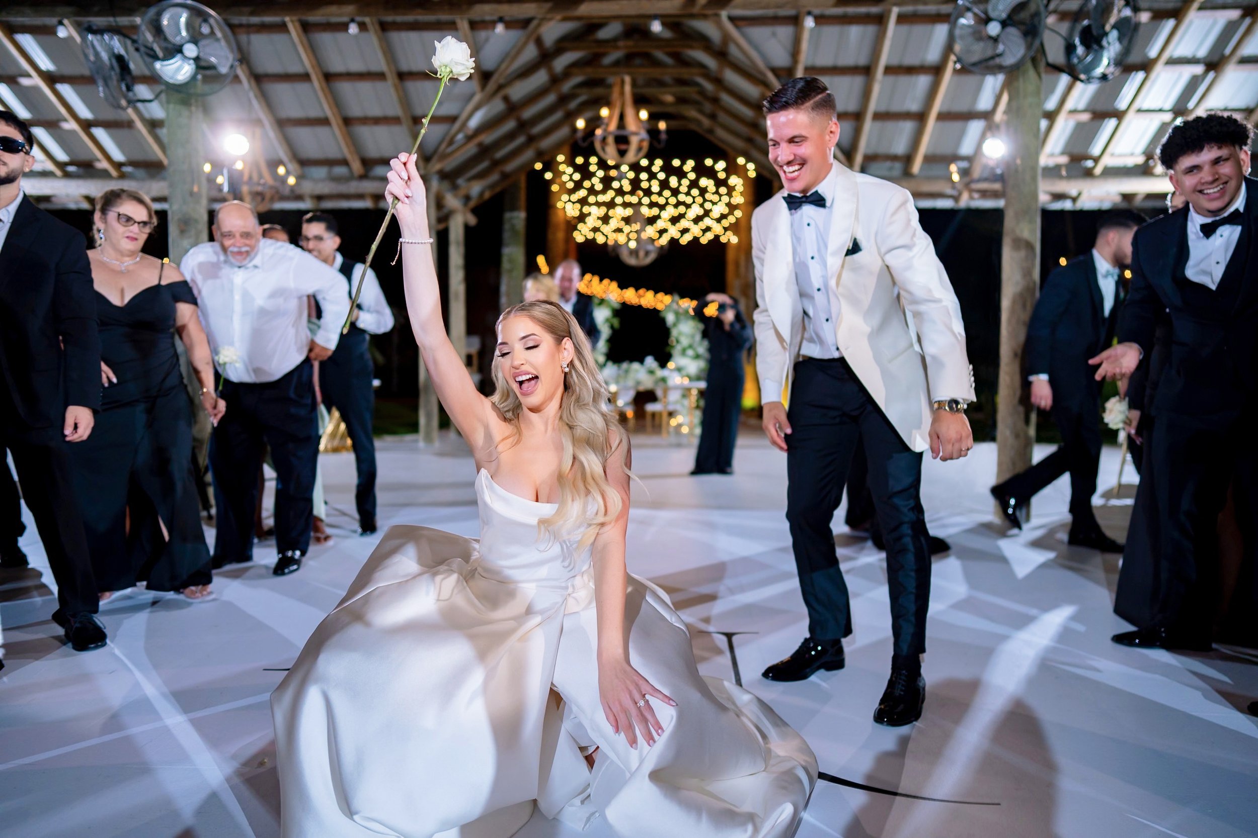 Bride in wedding dress kneeling and celebrating with a flower in her hand at a wedding reception, surrounded by guests dancing in a decorated venue with string lights.
