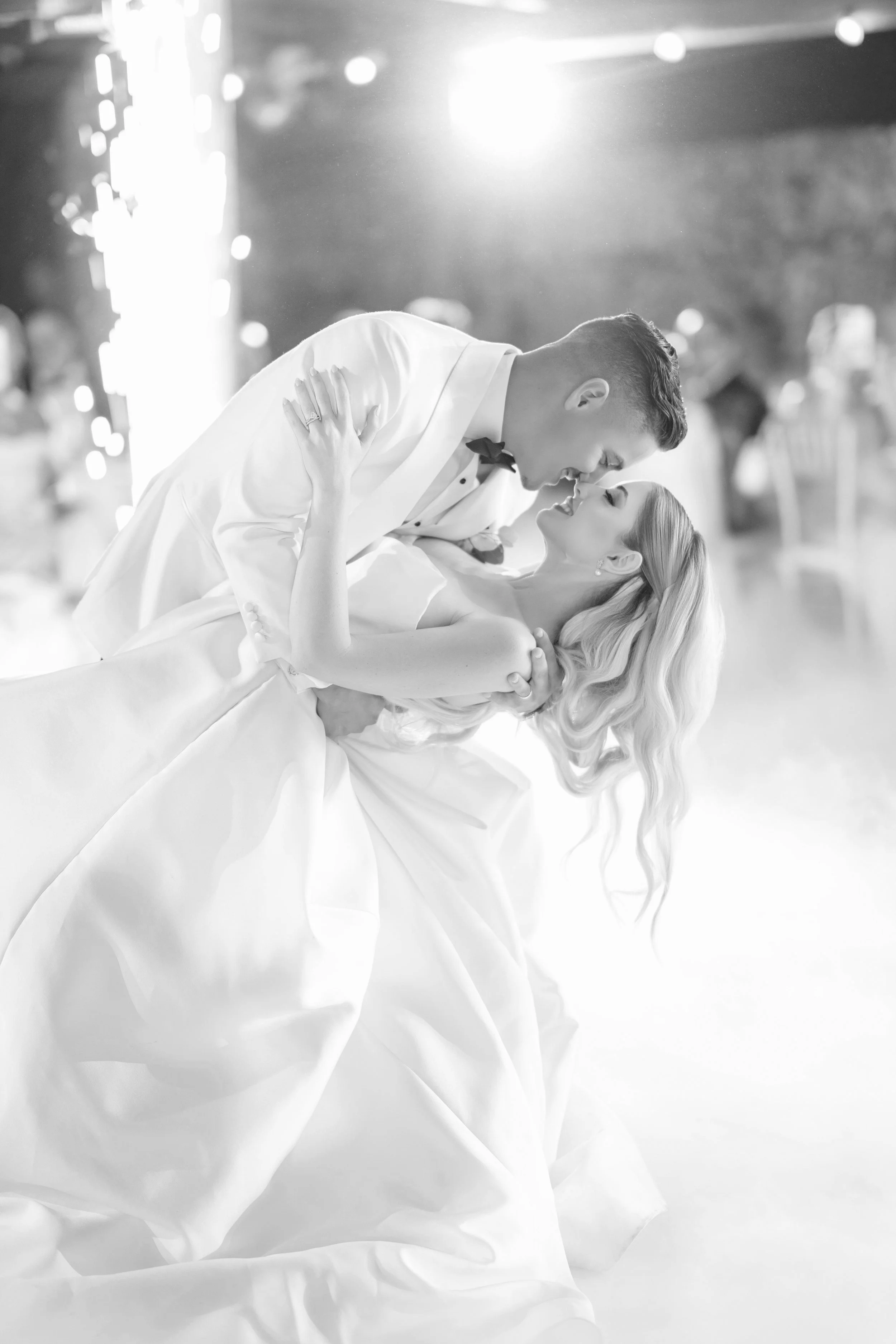 A black-and-white photo of a bride and groom dancing closely at their wedding reception, with the groom dipping the bride; the bride wears a white wedding gown and the groom a white tuxedo with a bowtie.