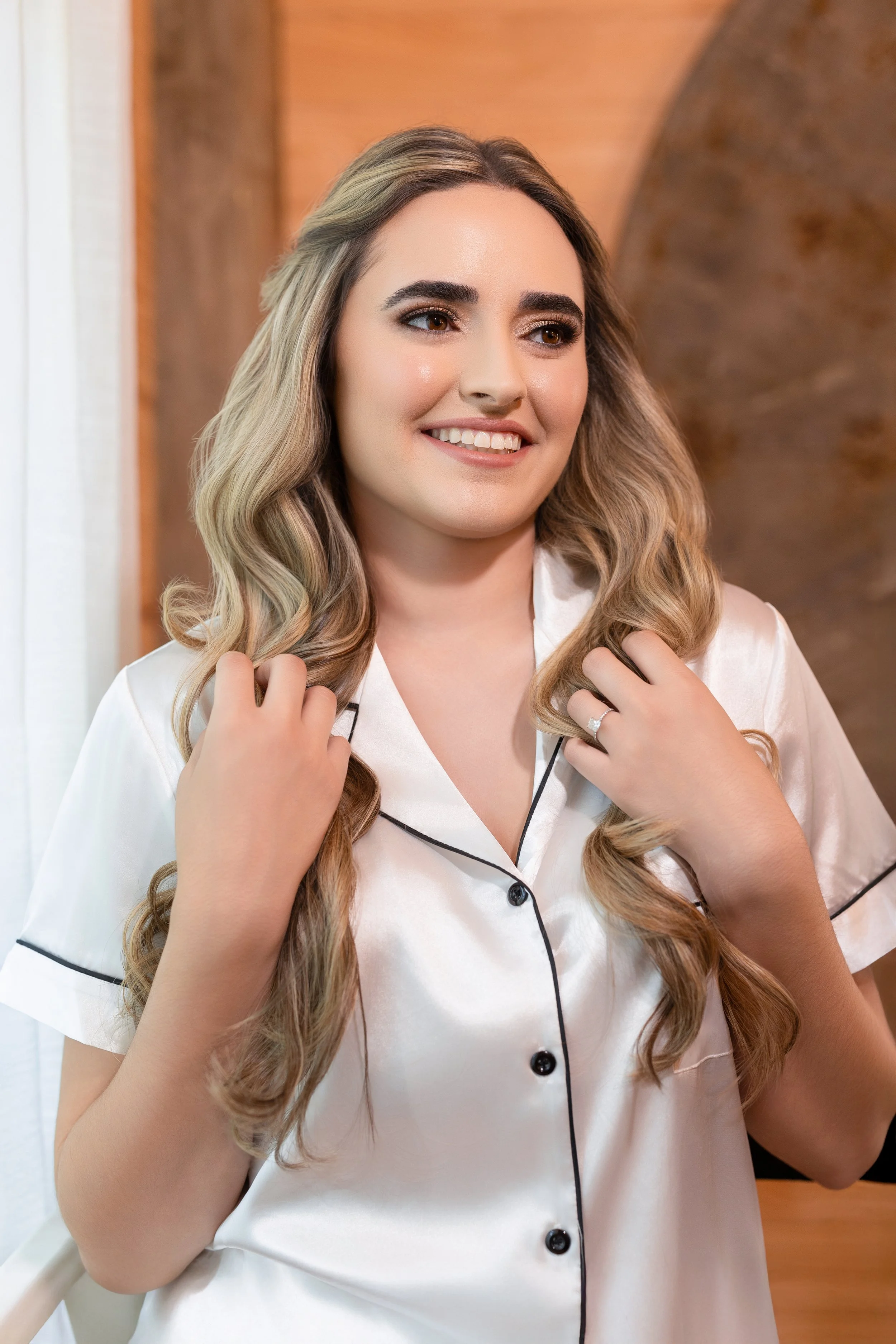 A woman with long, wavy blonde hair, wearing a white satin pajama top with black piping, smiling and adjusting her hair with both hands.