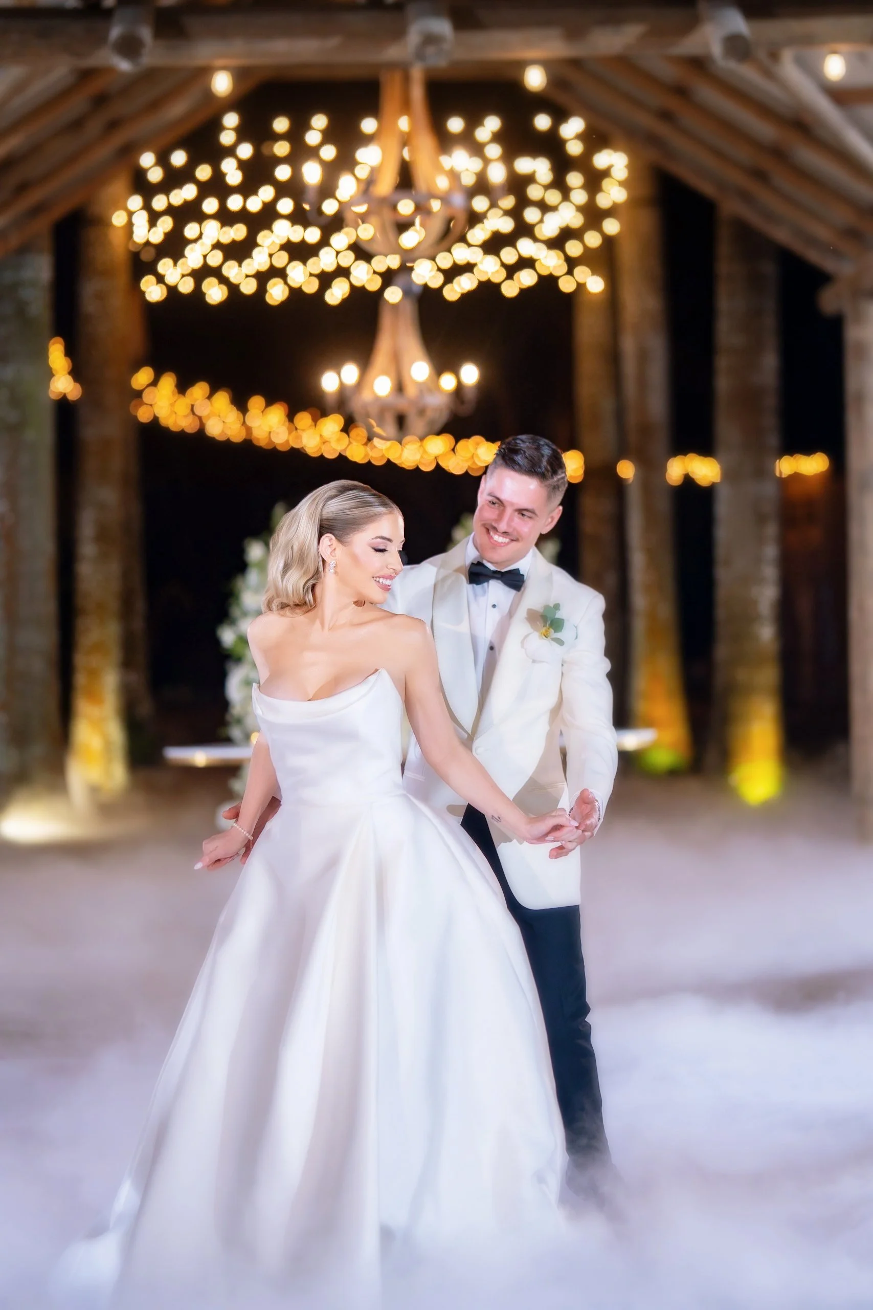 A bride and groom dancing at their wedding reception with warm lights and chandelier in the background.