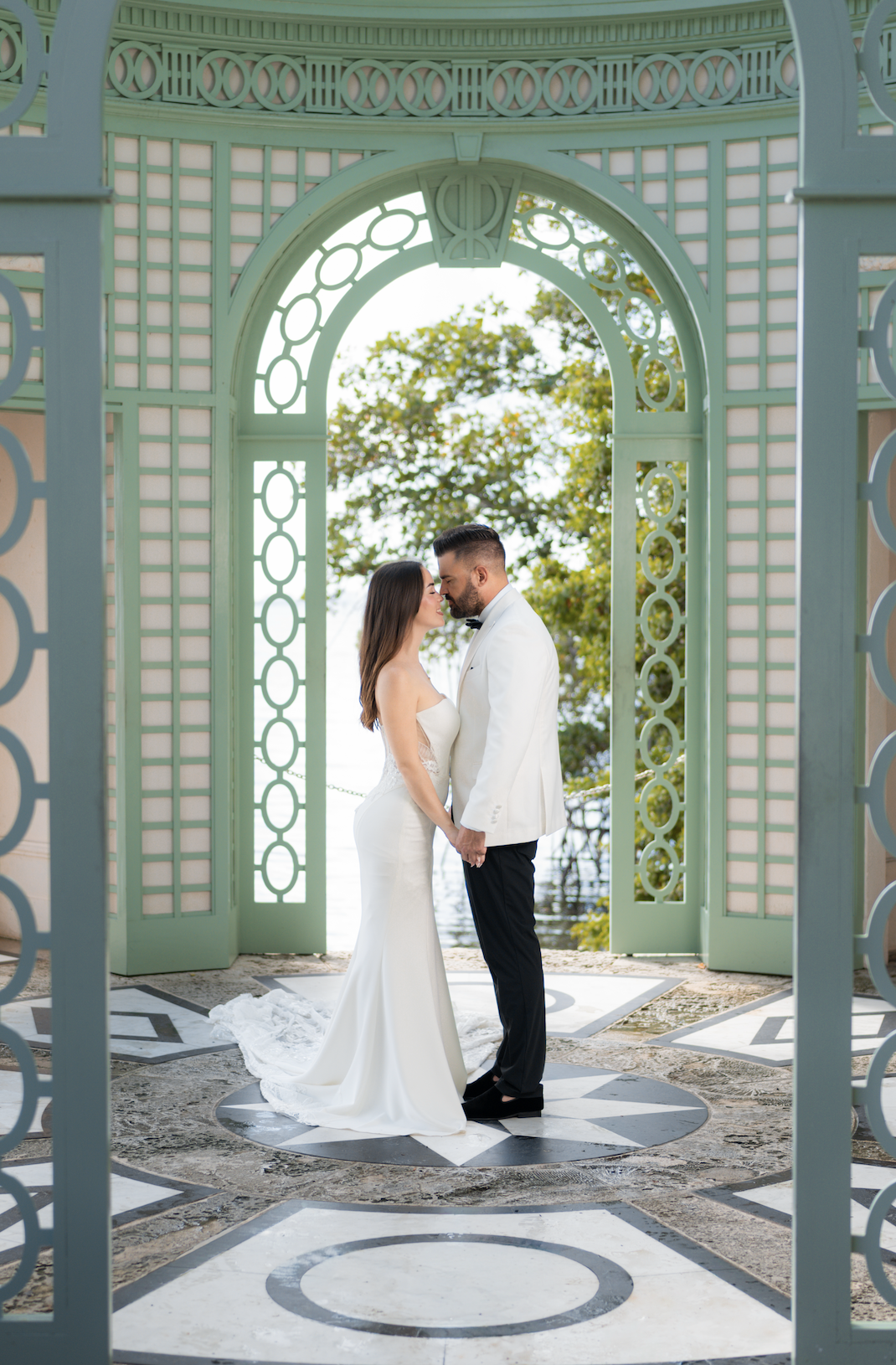 A bride and groom holding hands in a white wedding gown and tuxedo, standing close with foreheads touching inside a decorative, open structure with a scenic water and trees backdrop.