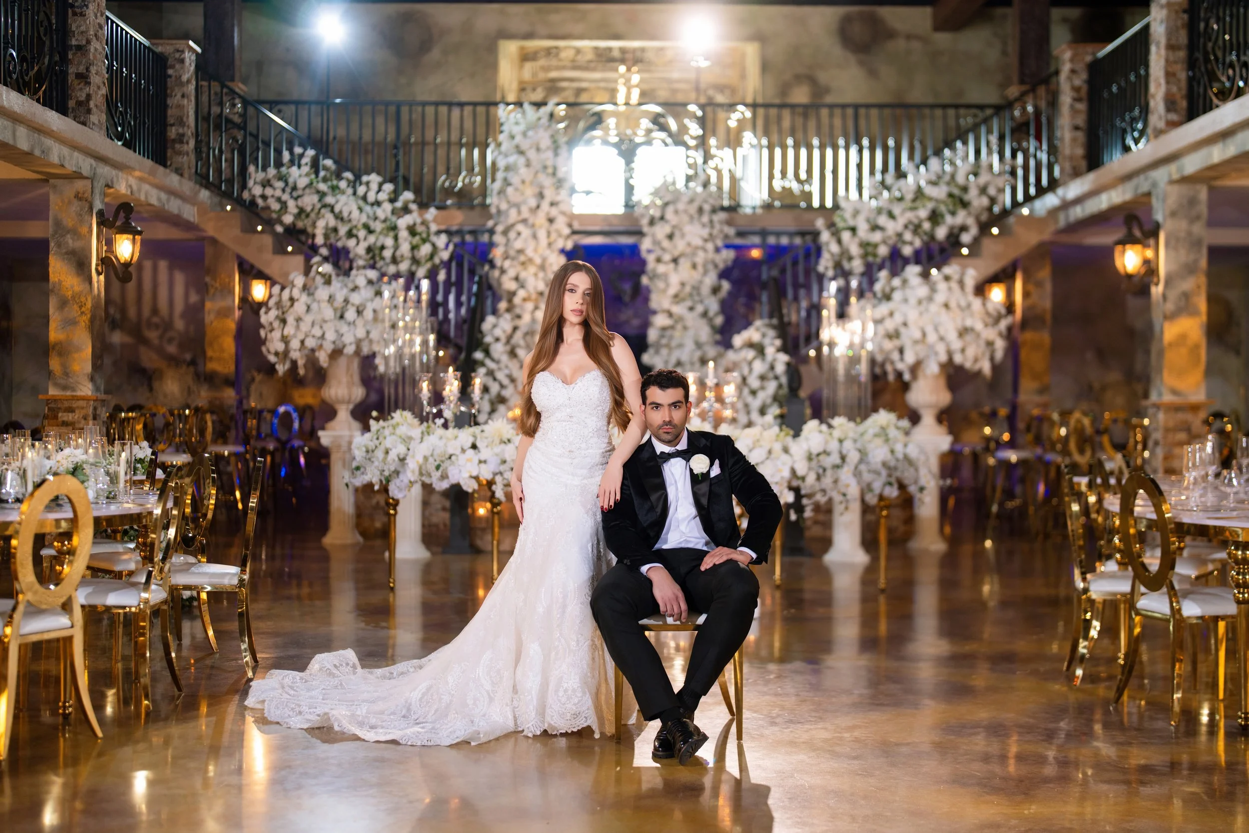 A bride and groom in a wedding venue decorated with white flowers, ornate gold chairs, and elegant lighting, with the bride standing and the groom sitting in front of a floral backdrop.