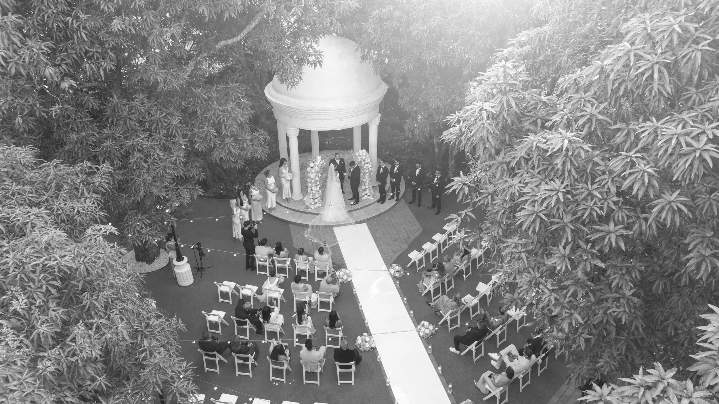 An outdoor wedding ceremony with a bride and groom standing in front of an officiant under a white, round gazebo. Guests are seated in white chairs on either side of a white aisle runner. The area is surrounded by large trees and string lights.