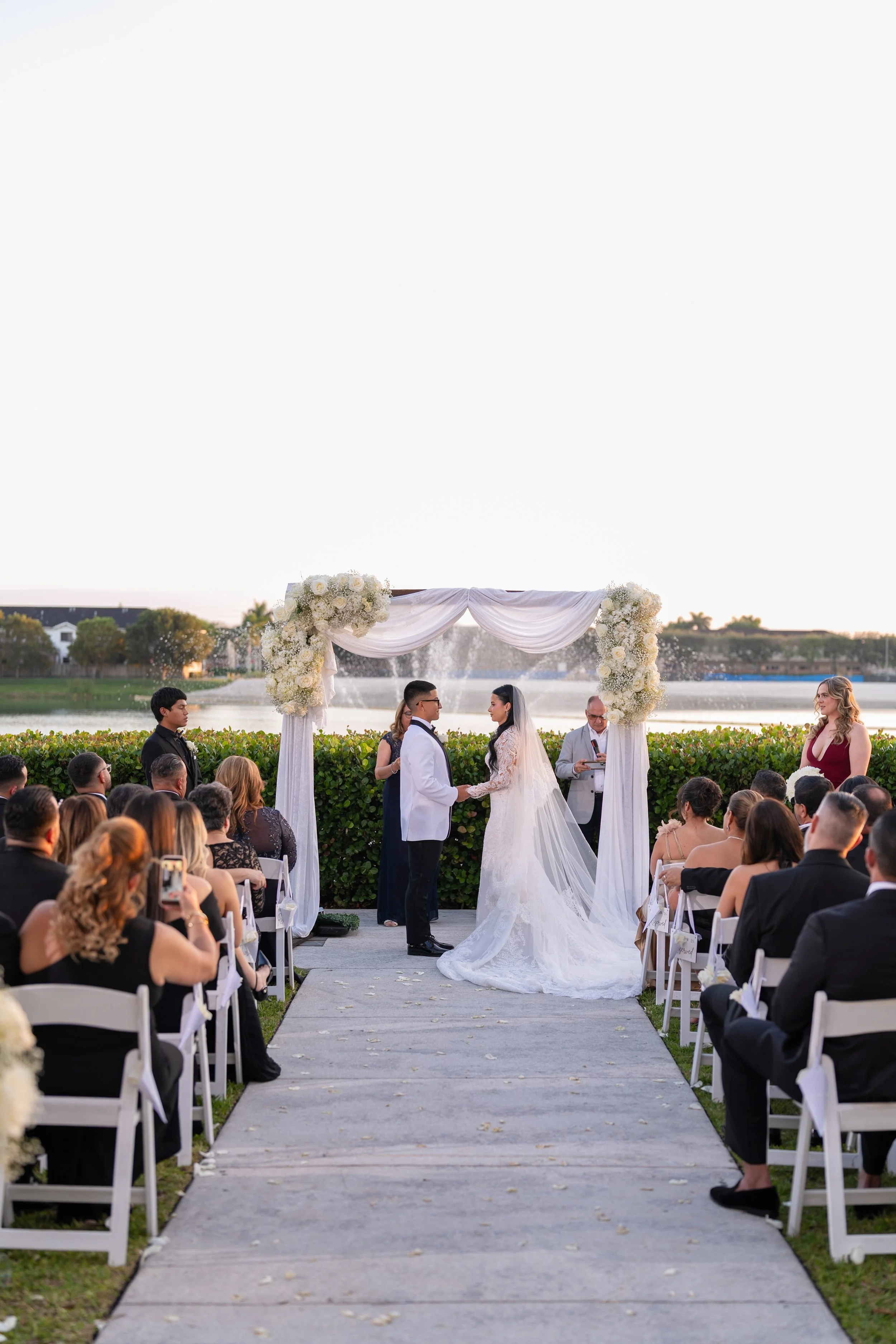 A wedding ceremony taking place outdoors by a lake, with the bride and groom holding hands under a decorated arch, surrounded by seated guests on either side.