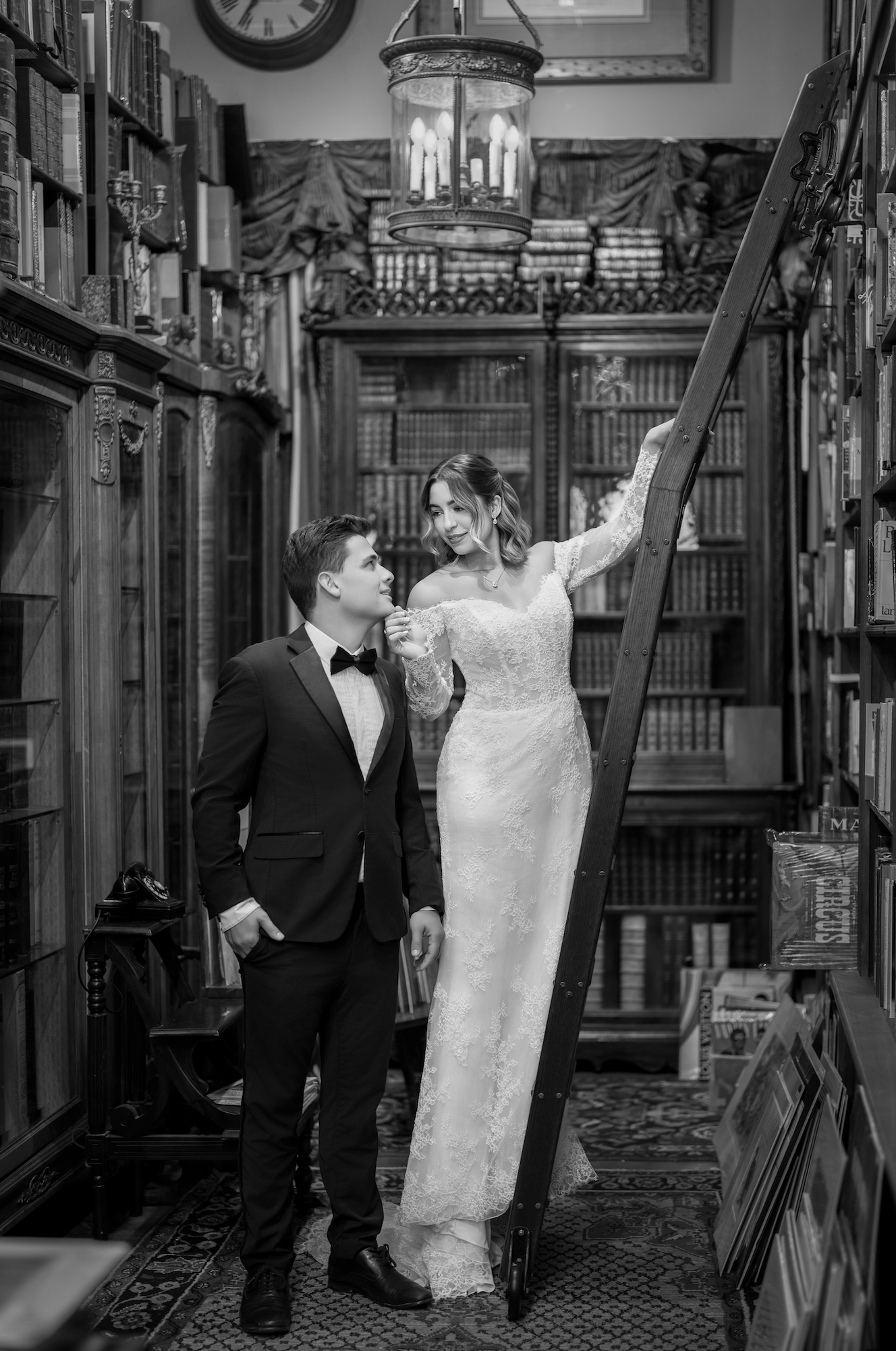 A man in a tuxedo and a woman in a wedding dress inside a library, with the woman standing on a ladder and looking at the man, who is looking up at her.