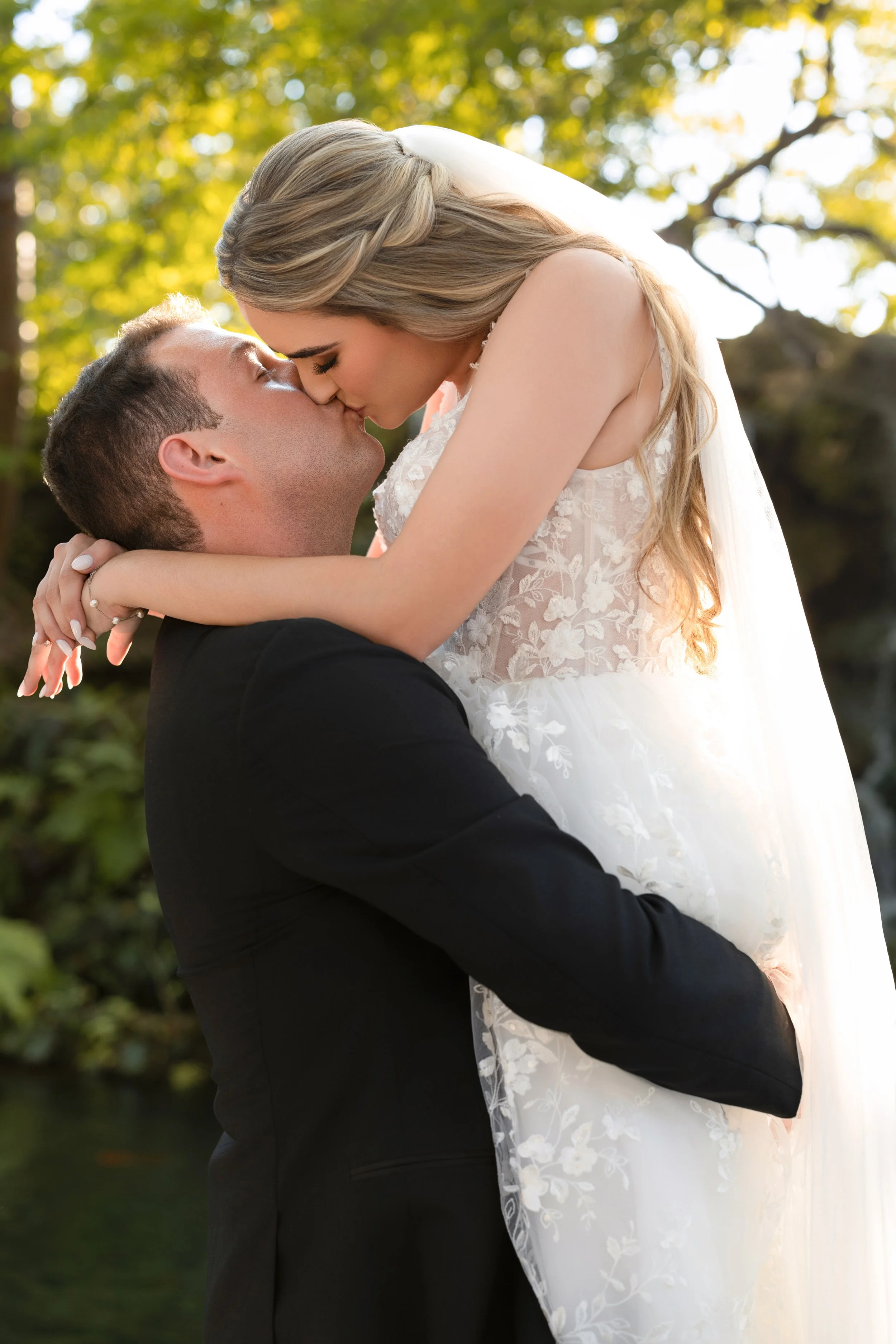 A newlywed couple sharing a kiss outdoors during sunset, with the man lifting the woman in his arms.