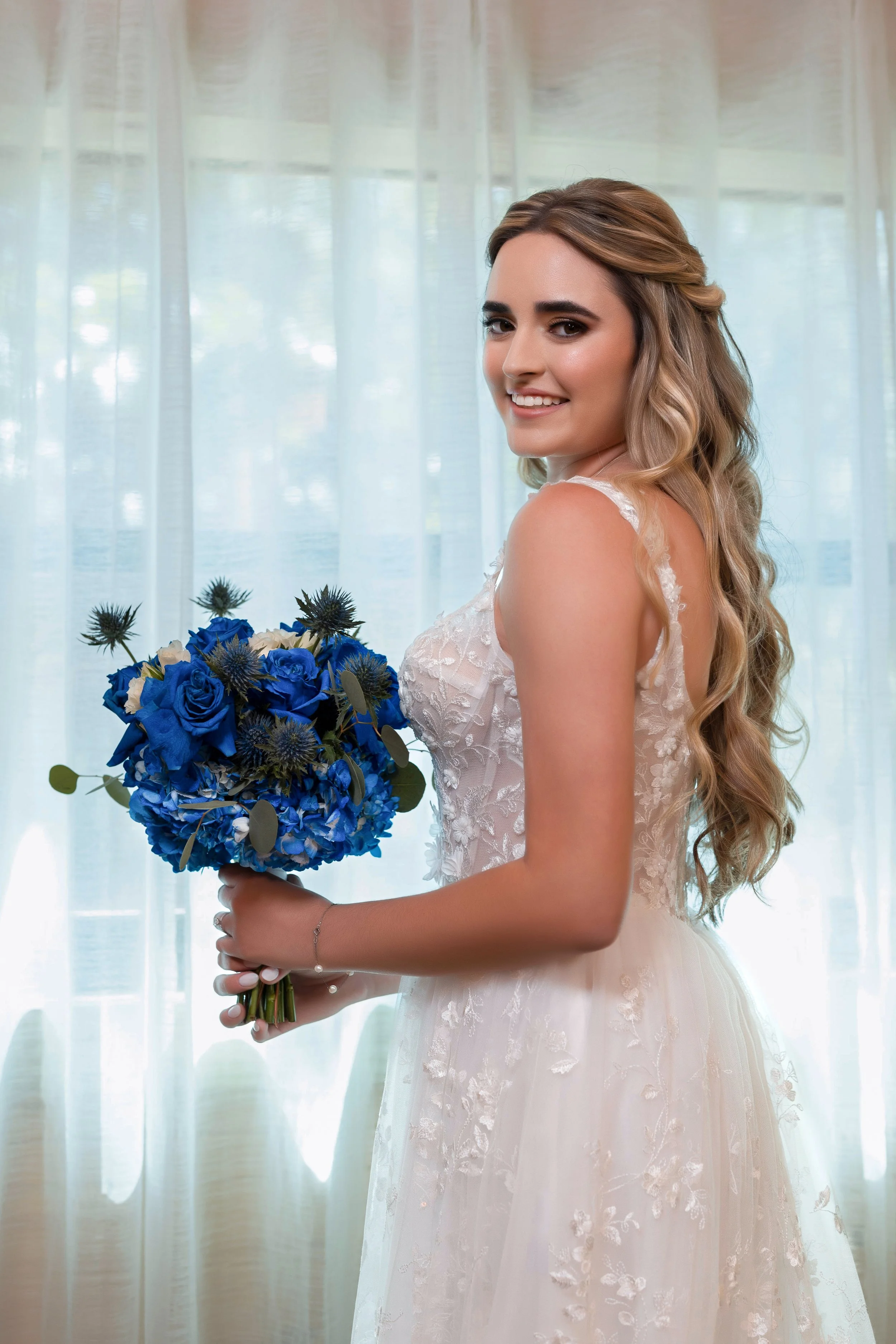 A smiling bride with wavy blonde hair in a white lace wedding dress holding a bouquet of blue flowers, standing in front of sheer curtains.