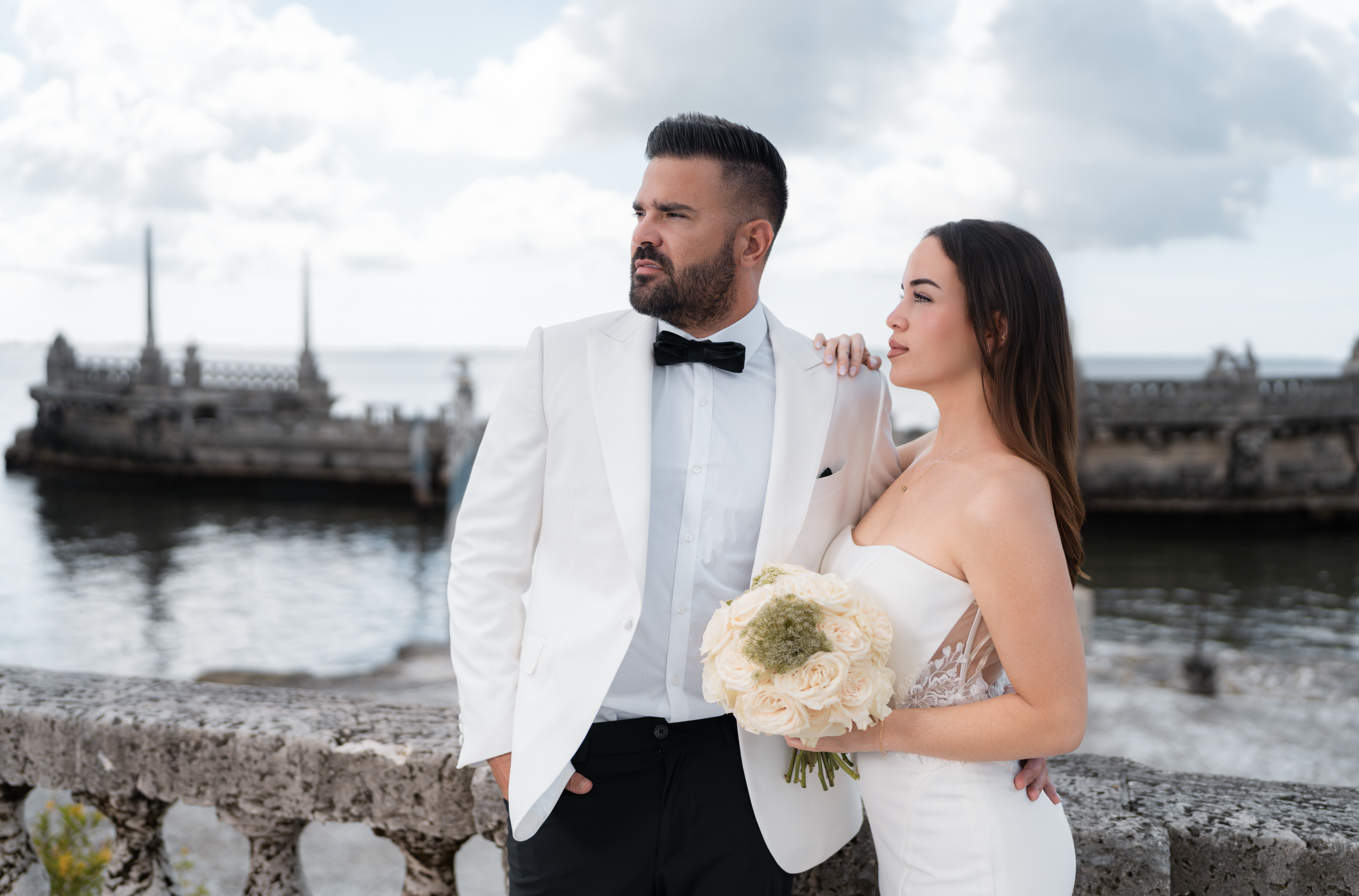 A groom in a white tuxedo and black bowtie standing by a bride in a strapless white wedding dress holding a bouquet of white roses at a waterfront.
