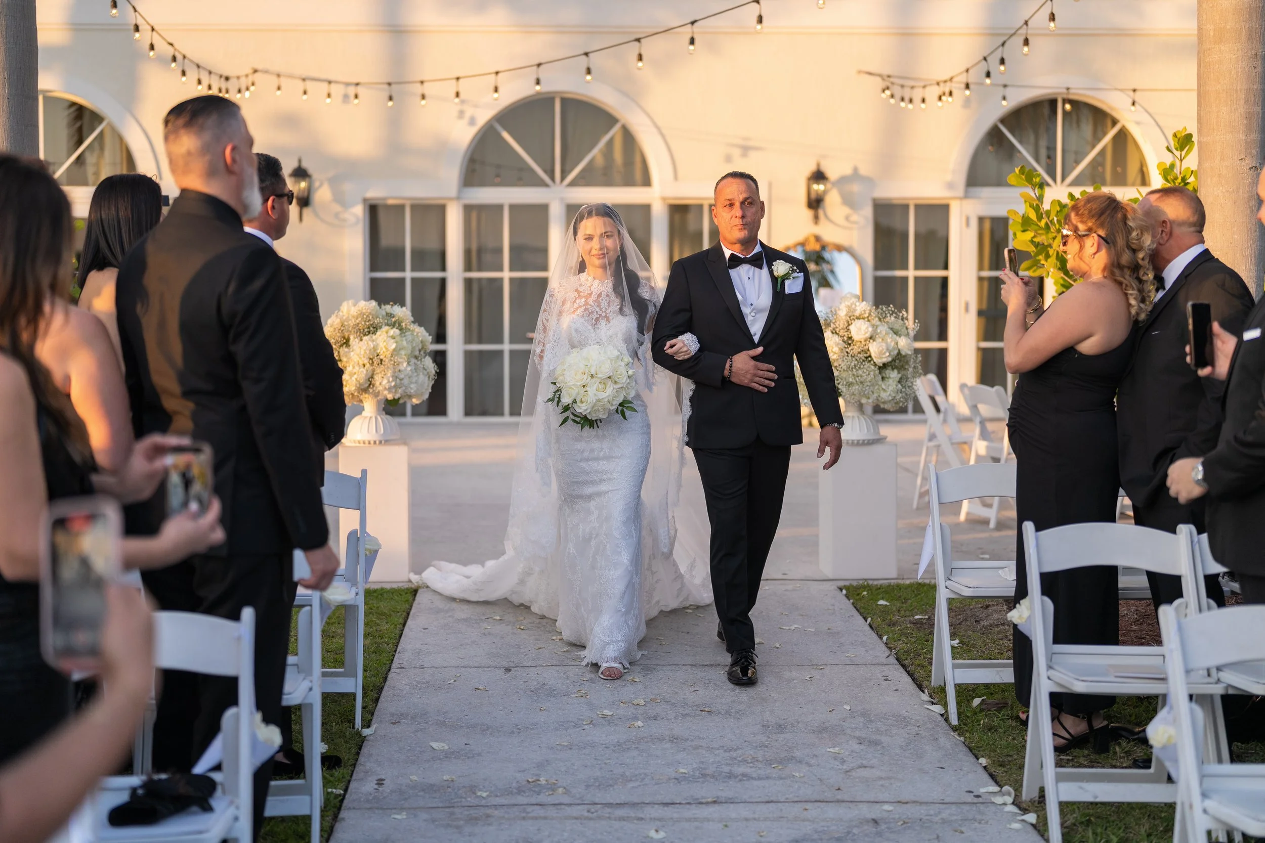 Bride walking down the aisle with her father at an outdoor wedding ceremony, guests standing and taking photos, decorated with white flowers and string lights.
