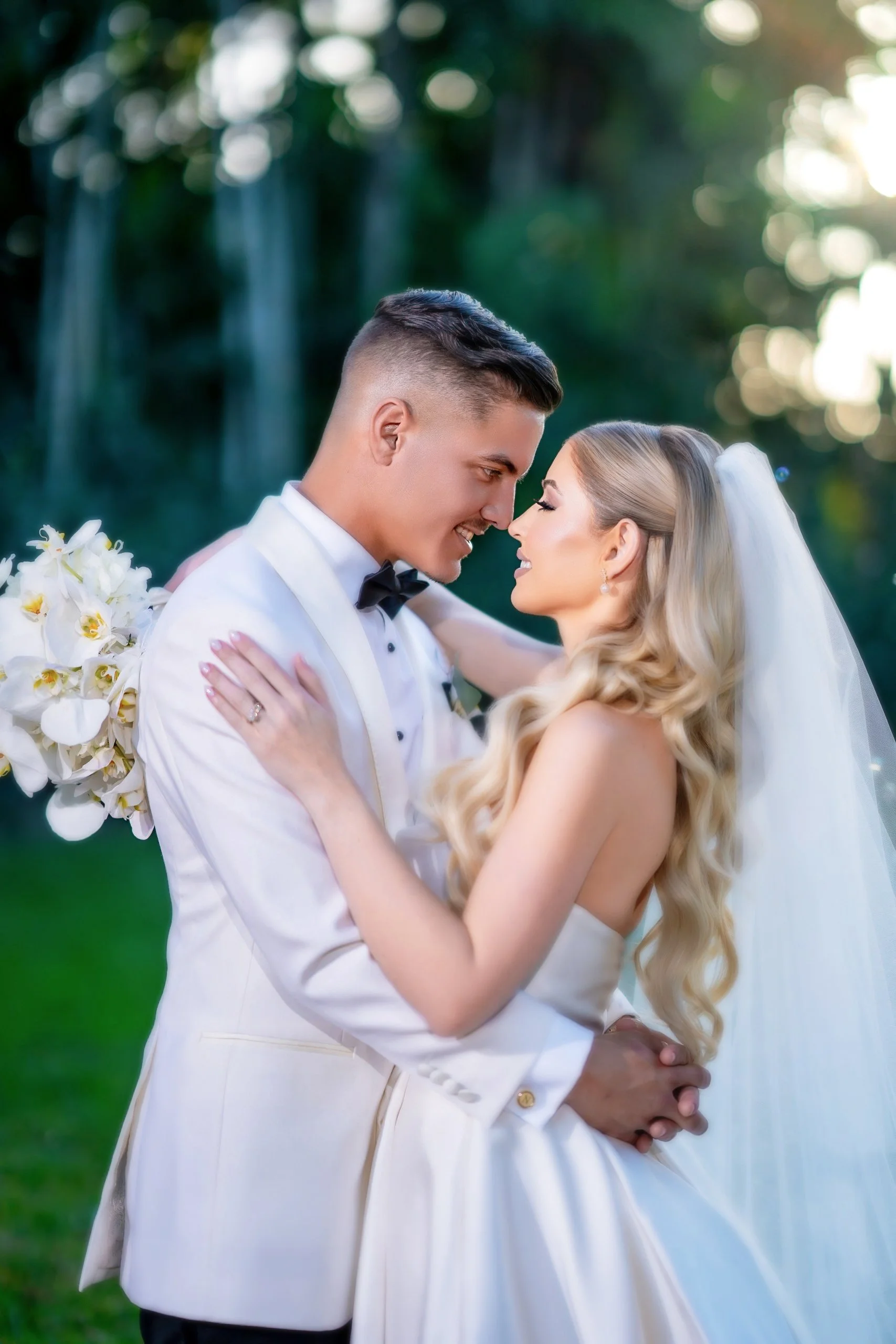 A bride and groom in wedding attire embracing outdoors, gazing into each other's eyes, with trees and sunlight in the background.