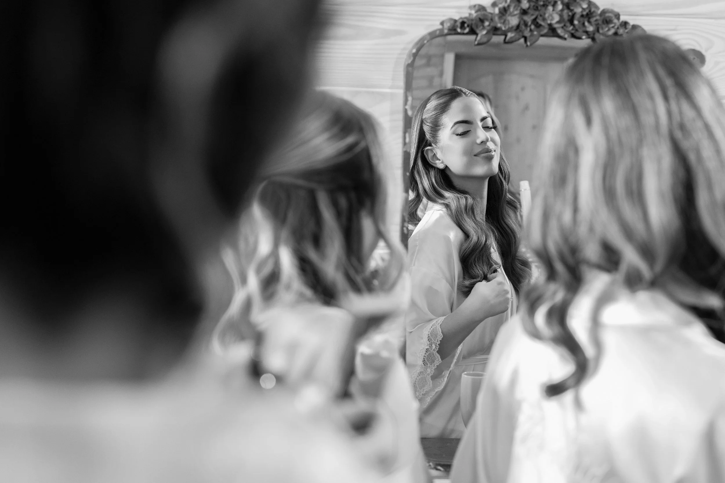 A woman with styled hair smiles while looking at her reflection in a mirror. She is wearing a satin robe with lace trim, surrounded by other women also preparing, in a cozy room with wooden walls.