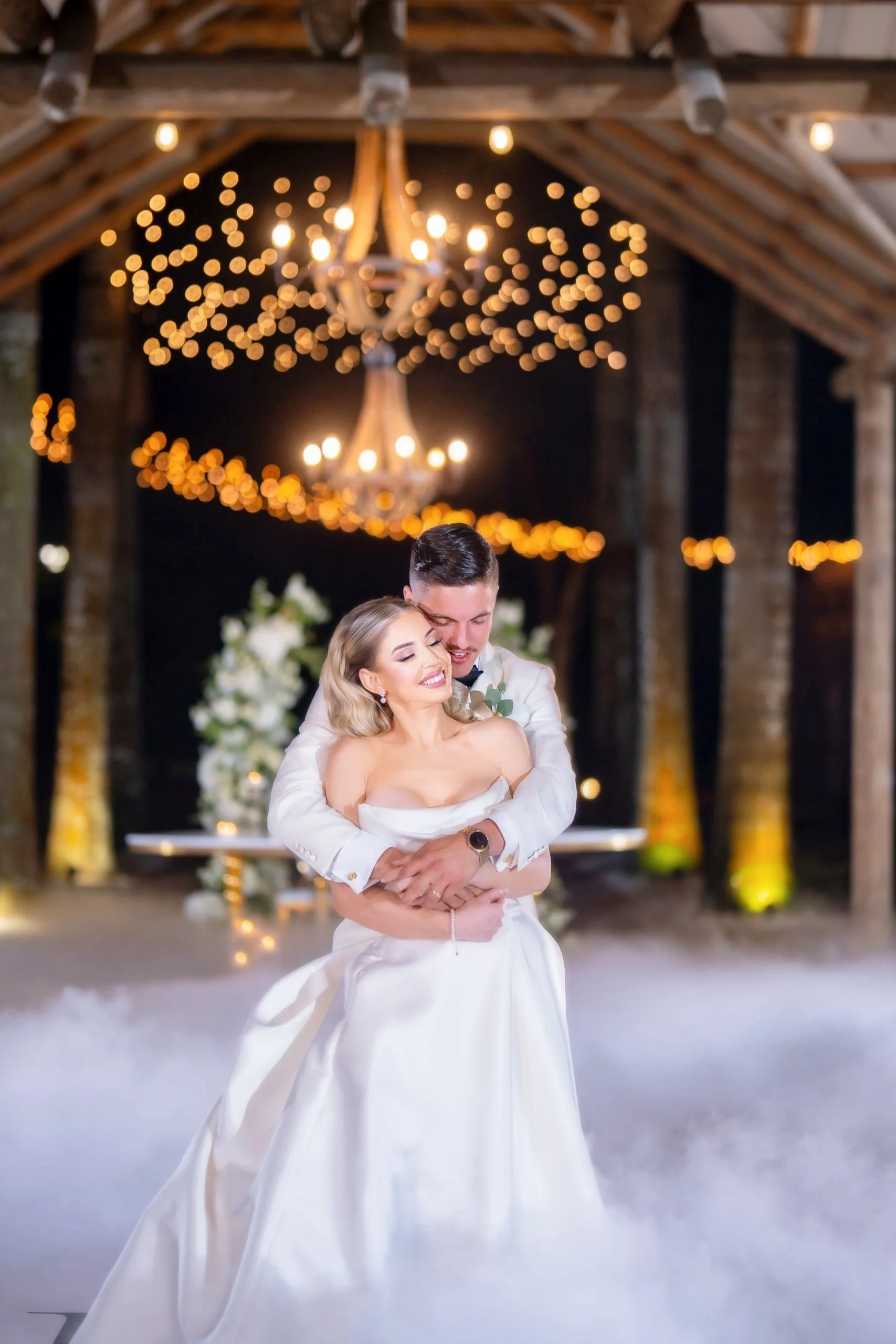 A bride and groom sharing a joyful dance at their wedding reception in a rustic barn with string lights and chandeliers in the background.