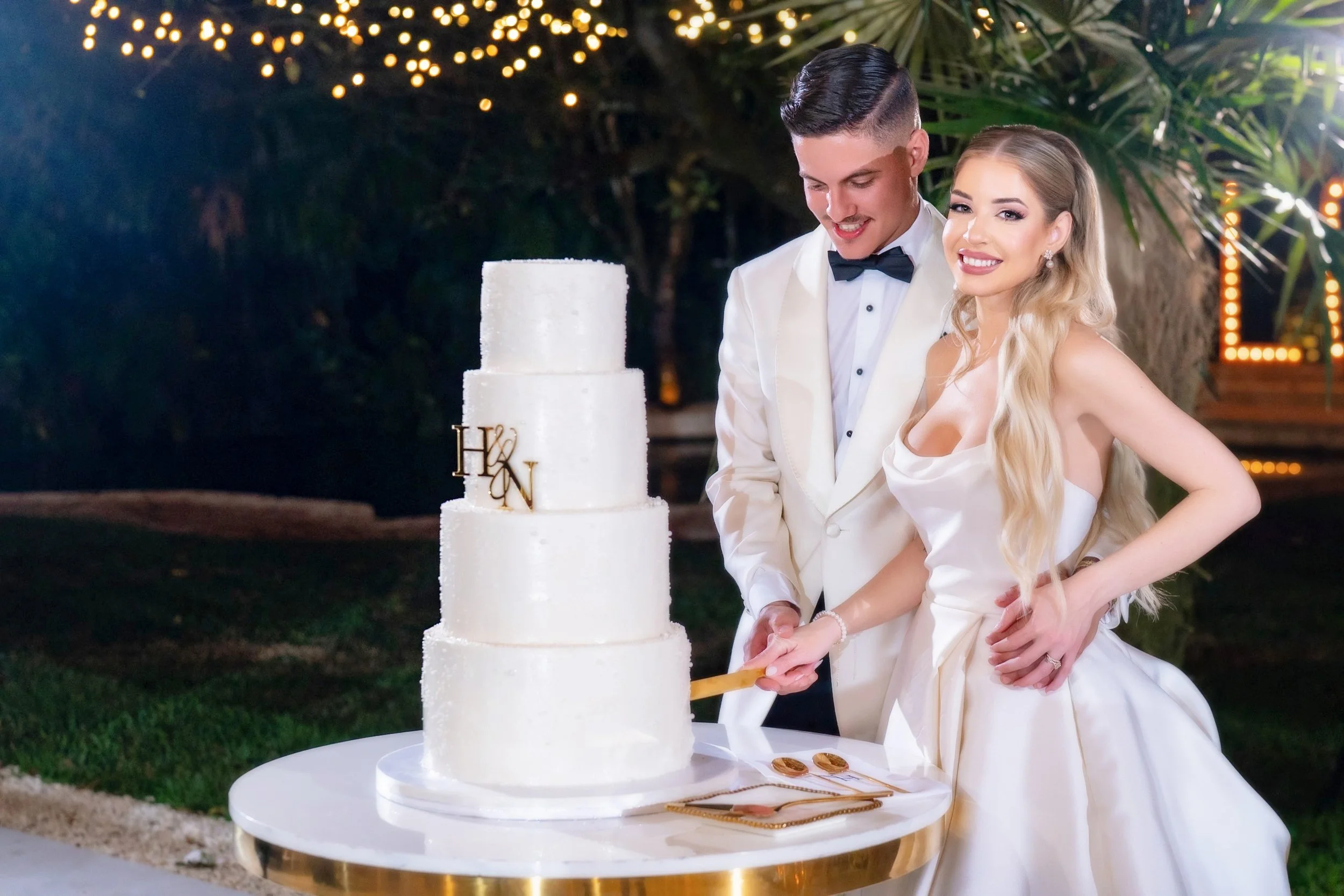 A newlywed couple in wedding attire cutting a tall white wedding cake with four tiers, outdoors at night decorated with string lights, with a woman smiling at the camera.