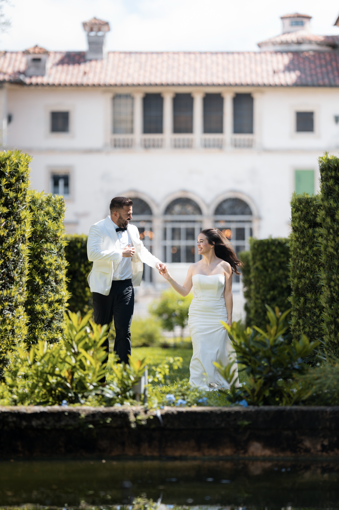 A bride and groom dressed in wedding attire are walking hand in hand in a lush garden, with a large historic building in the background.