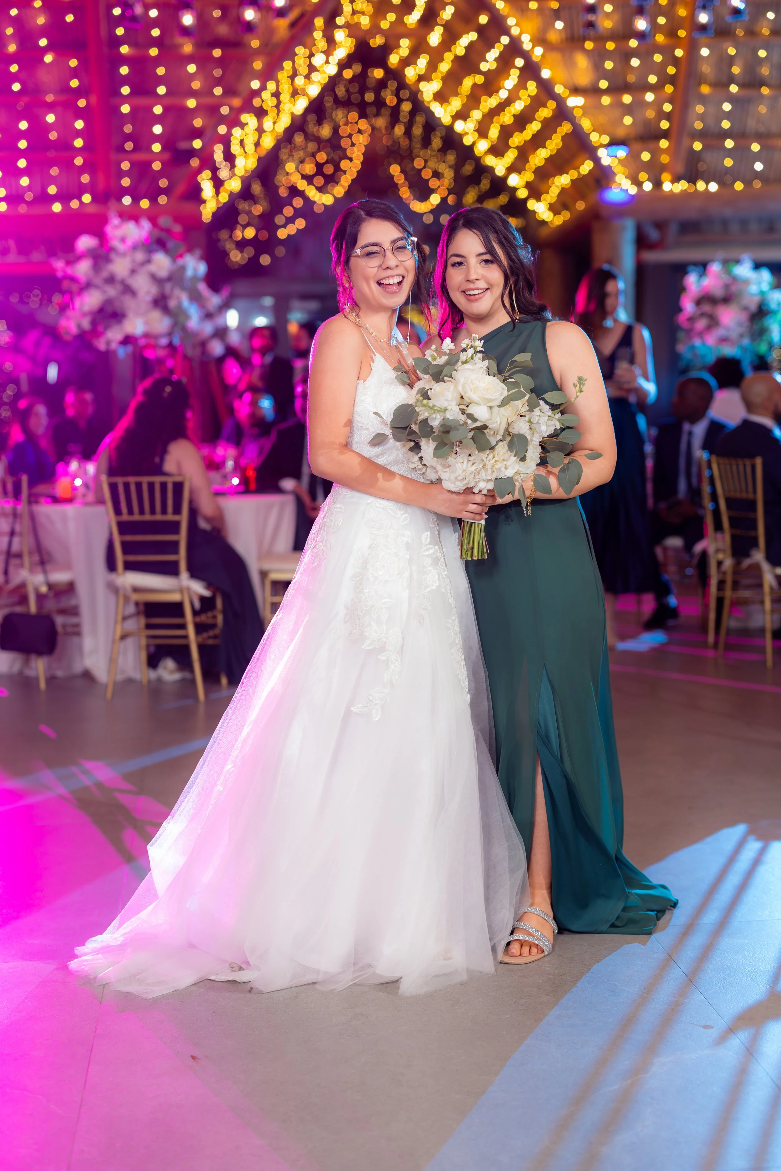 Two women smiling and posing together at a wedding reception, one in a white wedding gown holding a bouquet, and the other in a teal dress, in a decorated venue with string lights and floral arrangements.