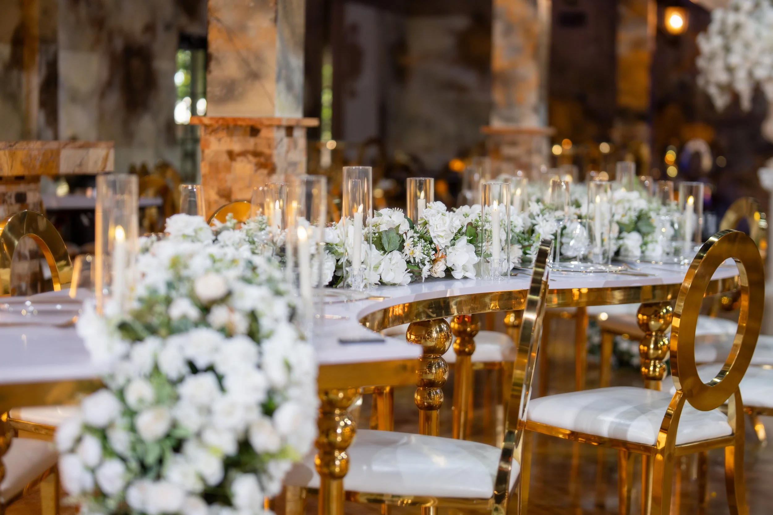 Elegant banquet table decorated with white flowers, glass candle holders, and gold chairs inside a rustic venue.