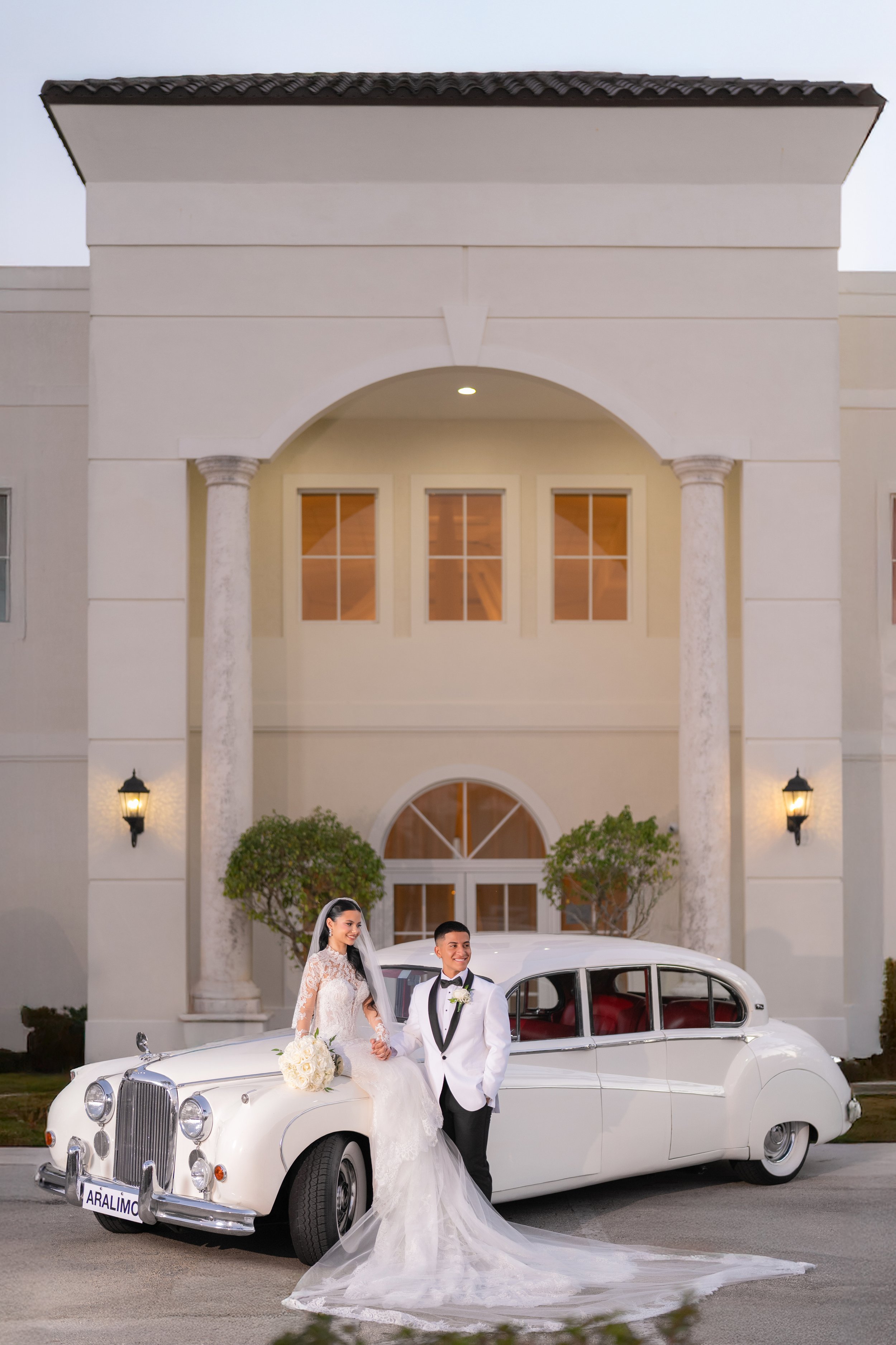 A bride and groom standing in front of a white vintage car parked outside a large elegant building. The bride is wearing a wedding gown with a long train, holding a bouquet, and the groom is dressed in a tuxedo with a bow tie.
