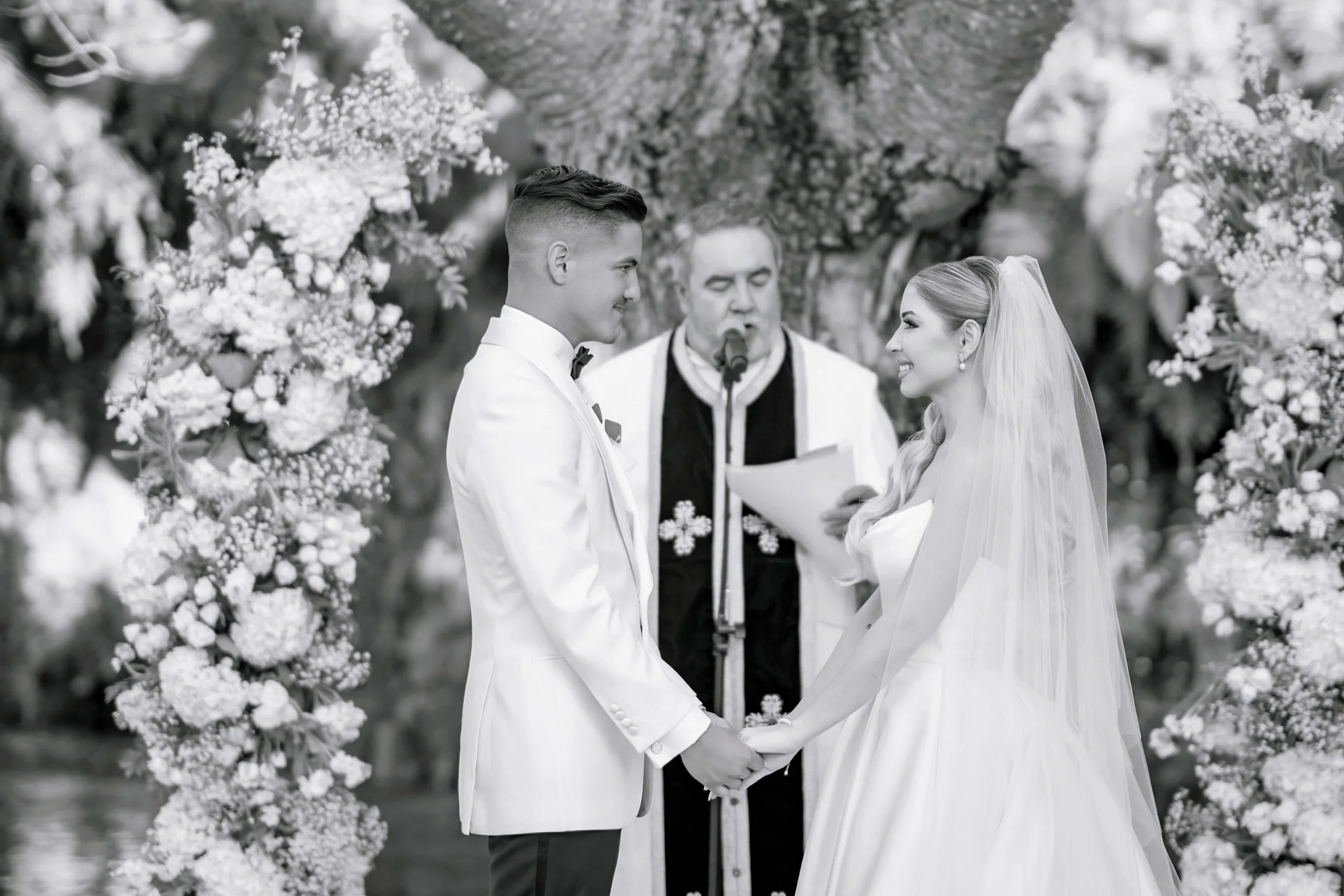 A black-and-white photo of a wedding ceremony with a bride and groom holding hands, standing in front of an officiant, surrounded by floral decorations.