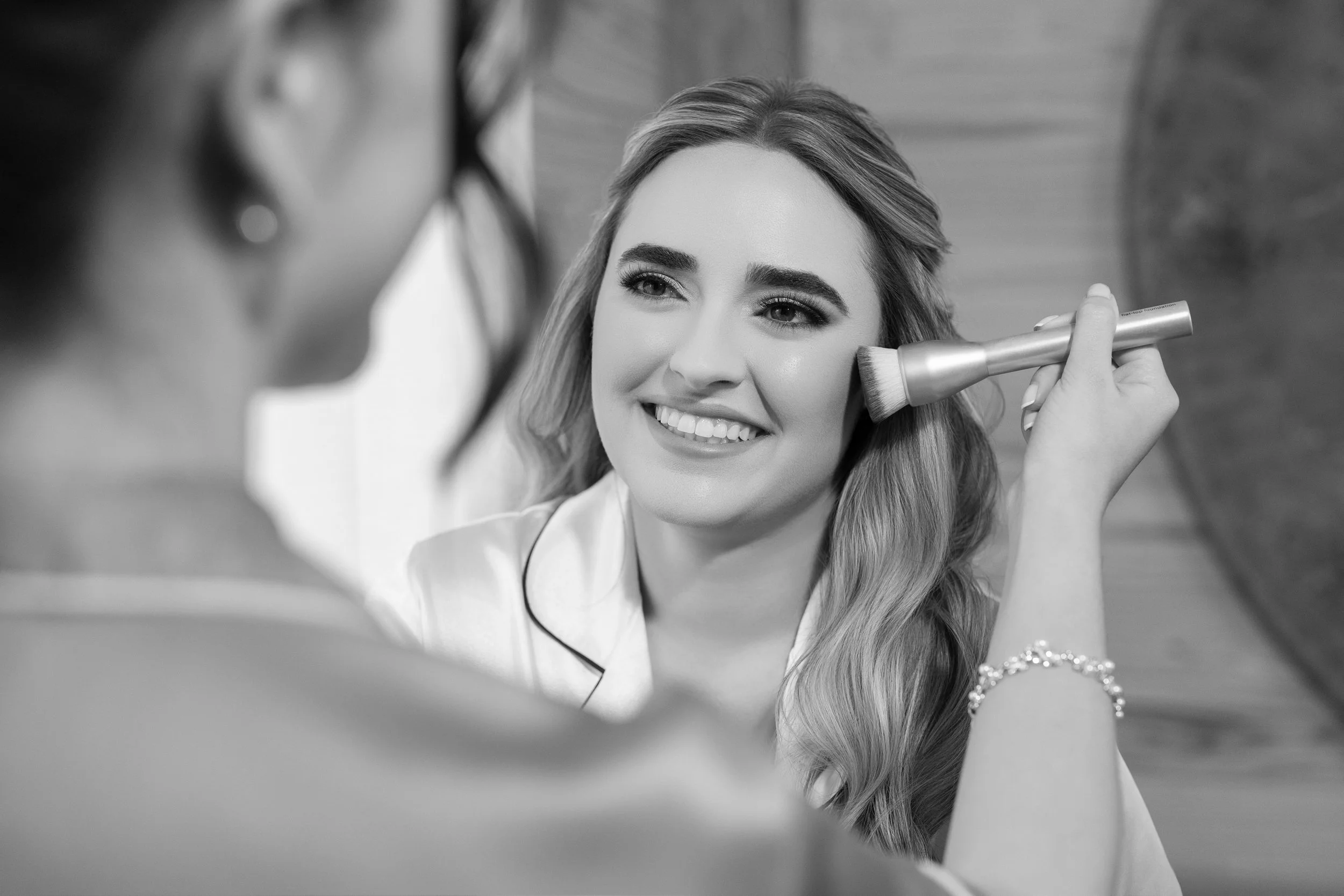 A woman smiling while having her makeup done, with another person applying makeup with a brush.