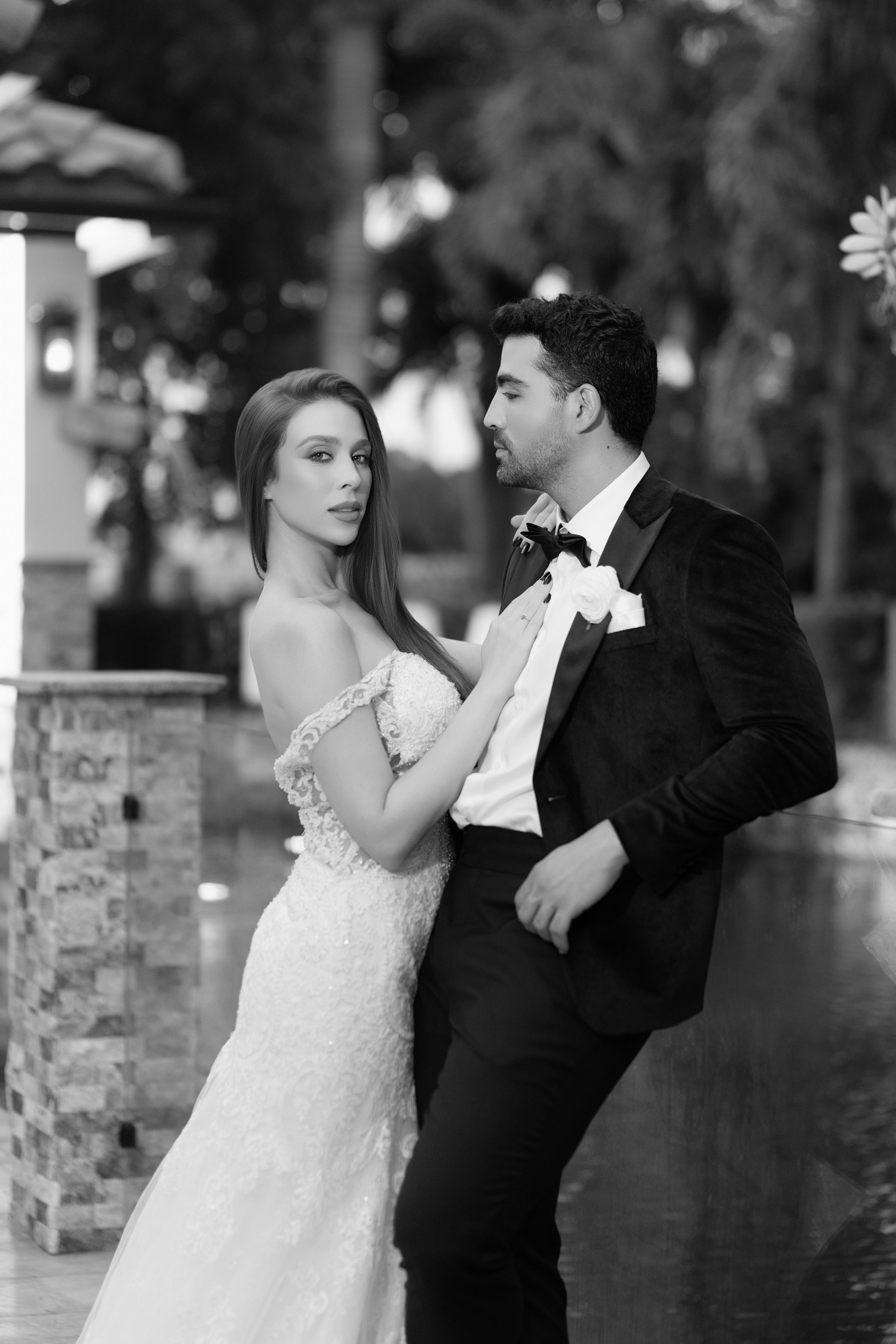 A black-and-white photo of a bride and groom posing together outdoors. The bride has long hair and wears a lace wedding dress, while the groom is in a tuxedo with a bow tie. They are close, with the bride's hand on the groom's chest, looking at the camera and each other.