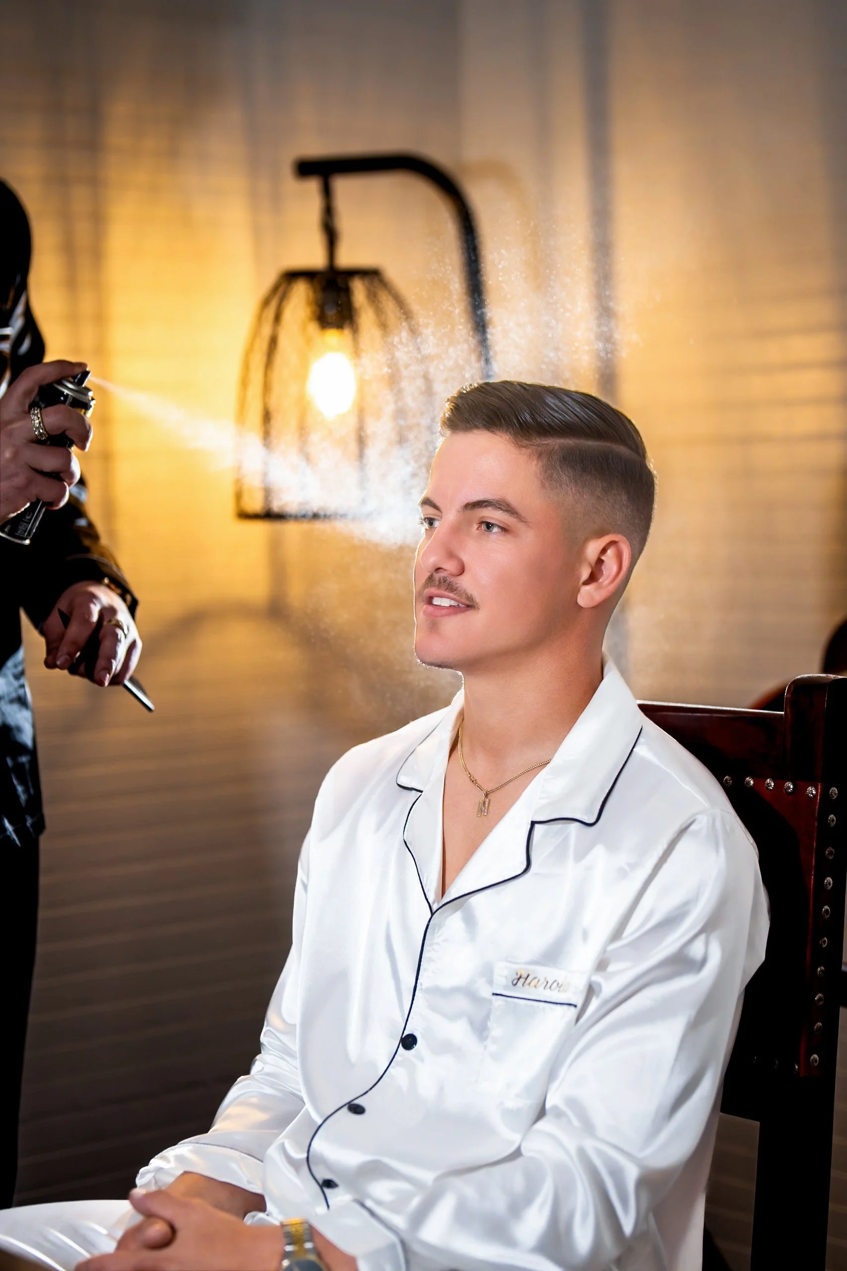 A young man in white pajamas is sitting while someone is spraying hair spray on his hair. The background has warm lighting with a hanging lamp and a brick wall.