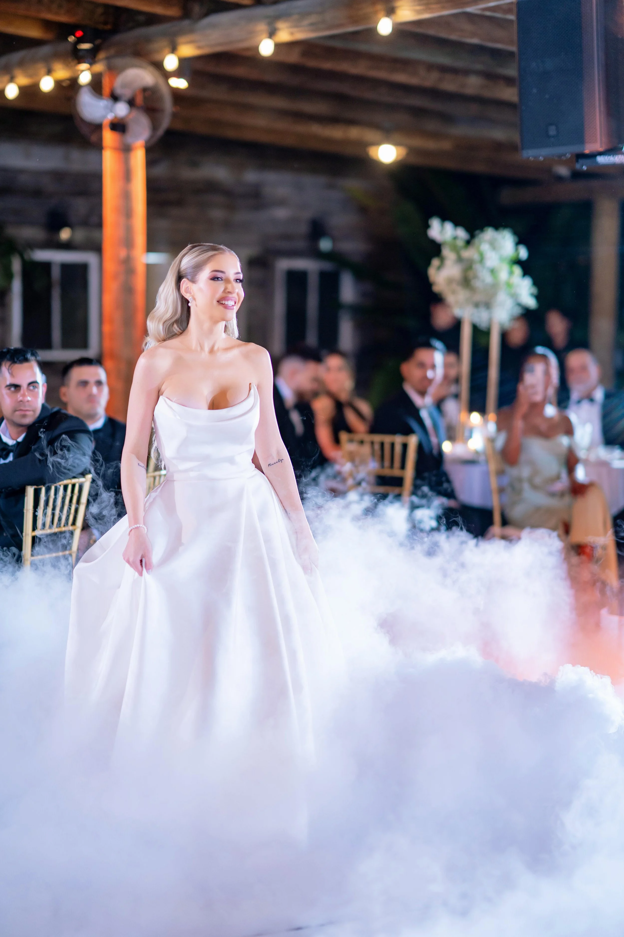 A bride in a white wedding dress walking through fog at her wedding reception, with guests seated at decorated tables in a rustic venue.