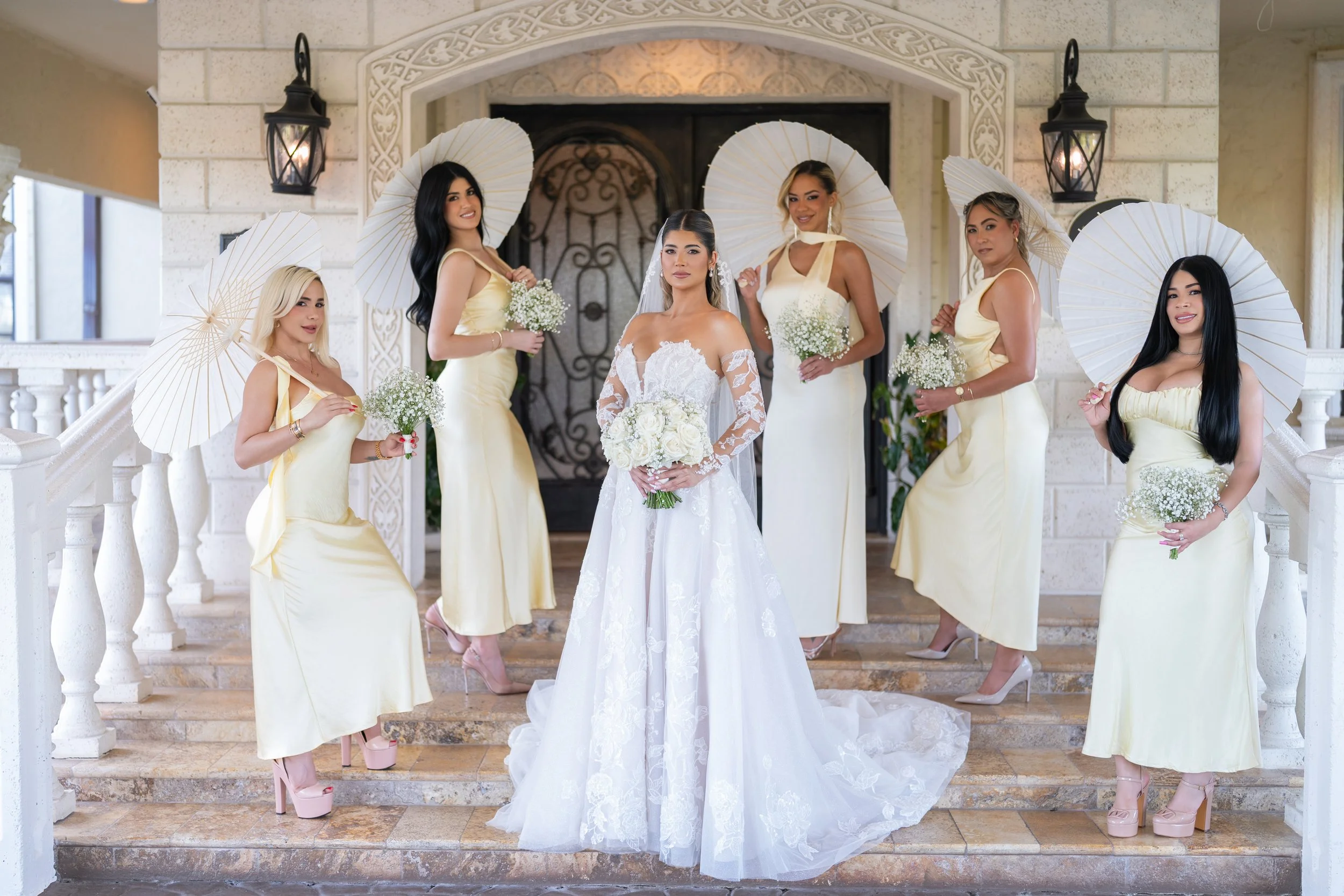 A bride in a white lace wedding gown with a veil holding a bouquet of white roses; she is surrounded by six bridesmaids dressed in matching long yellow satin dresses, each holding a white parasol and a small bouquet of baby's breath flowers, standing on a staircase in front of a decorative archway with lanterns.