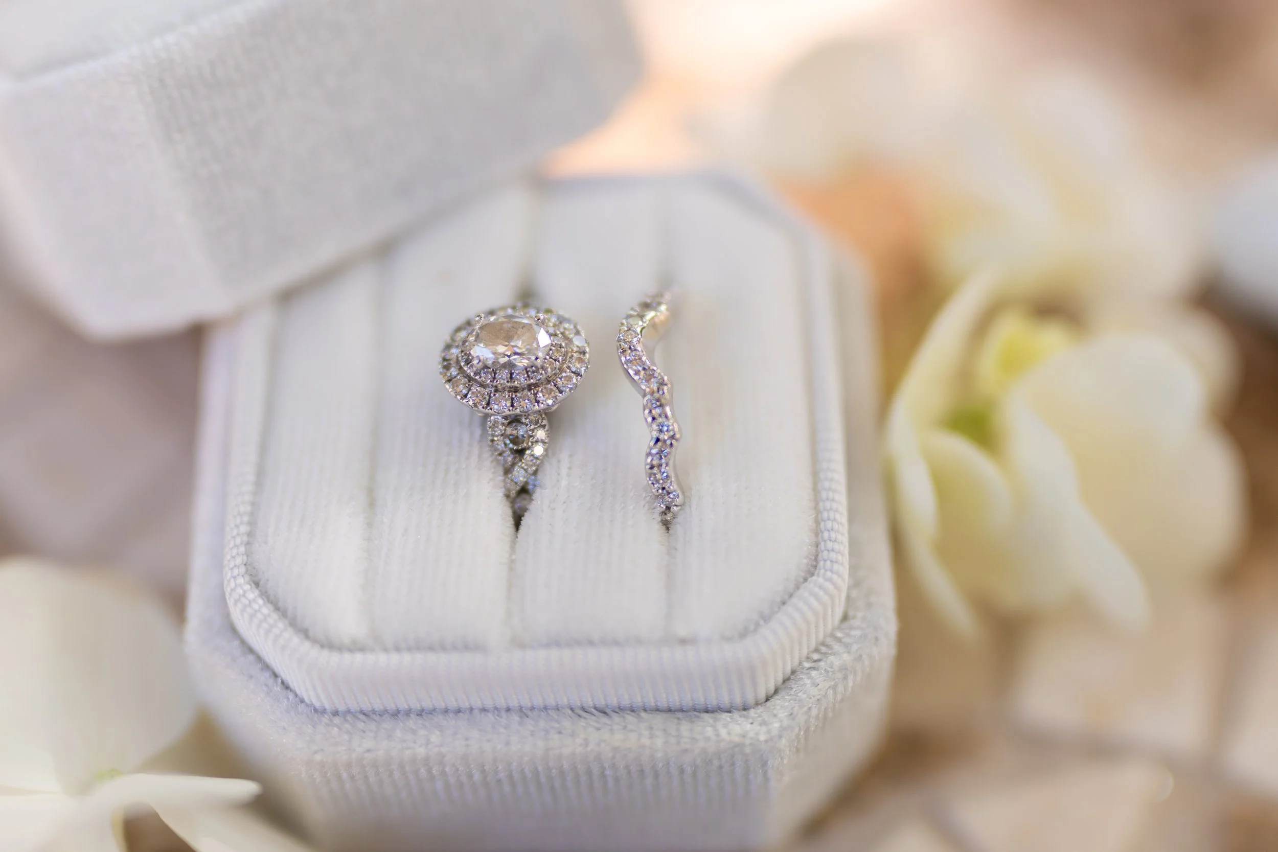 Close-up of a diamond engagement ring and matching wedding band in a white jewelry box, surrounded by white flowers.