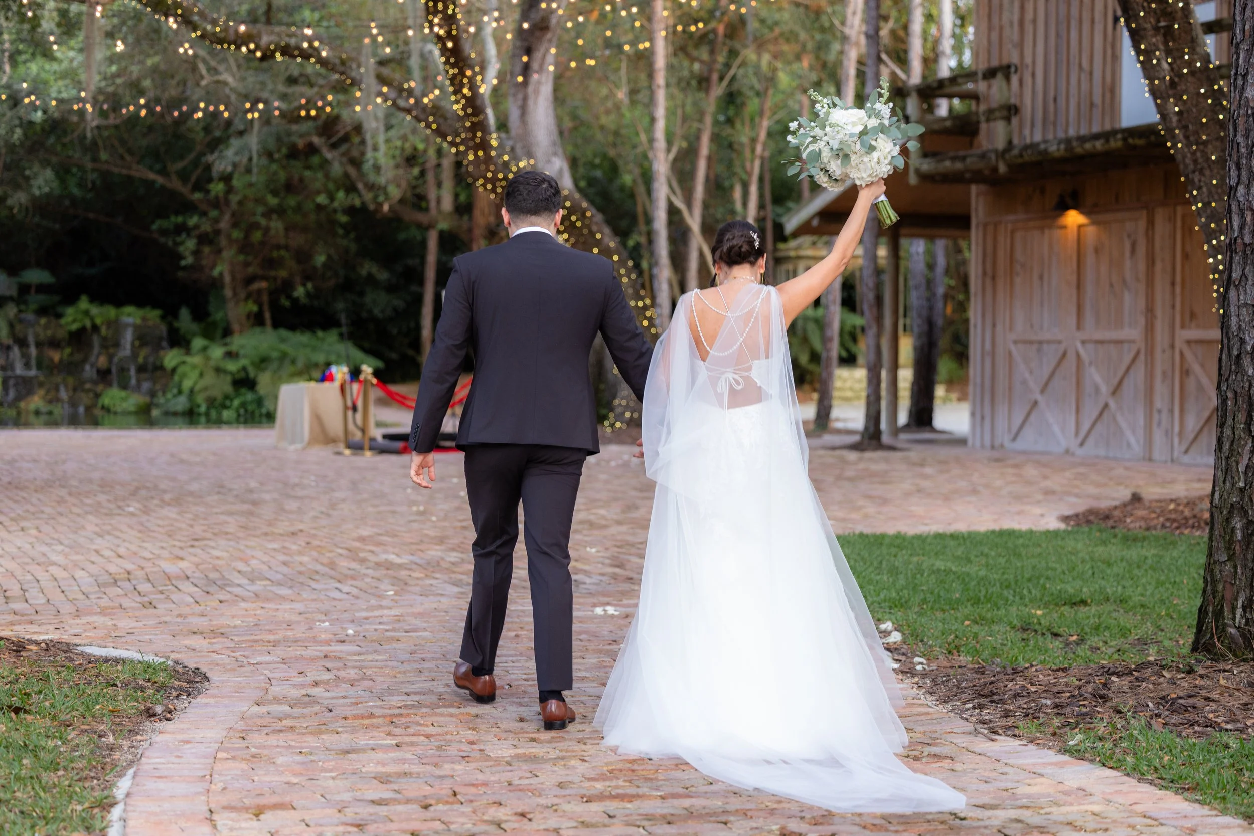 A bride and groom walking hold hands outdoors during their wedding. The bride is wearing a white wedding dress and veil, carrying a bouquet of white flowers, with her arm raised. The groom is dressed in a black suit. There are string lights hanging i