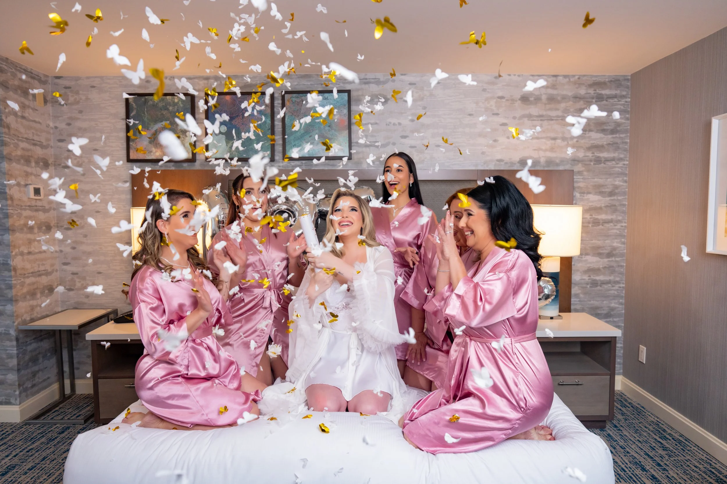 Joyful bride and bridesmaids celebrating in pink robes with confetti at HILTON TAMPA DOWNTOWN; fun wedding preparation photography by Star Visual Art, Tampa.