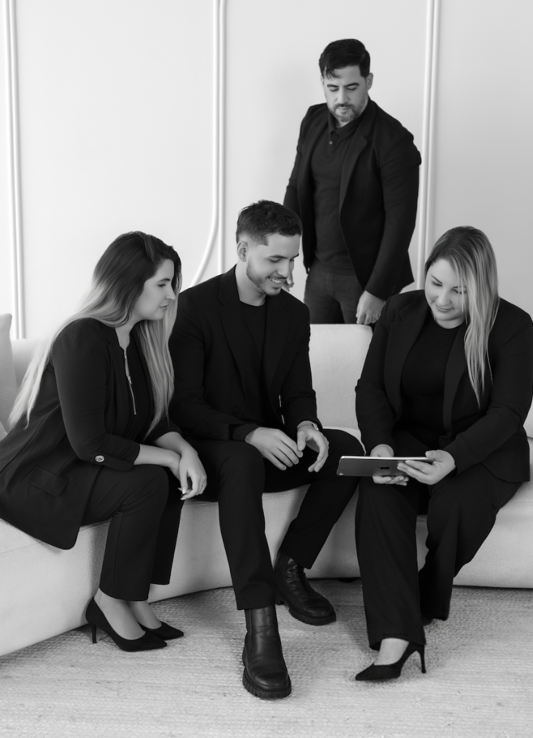 Four business professionals dressed in formal black suits sitting and standing in an office, looking at a tablet together.