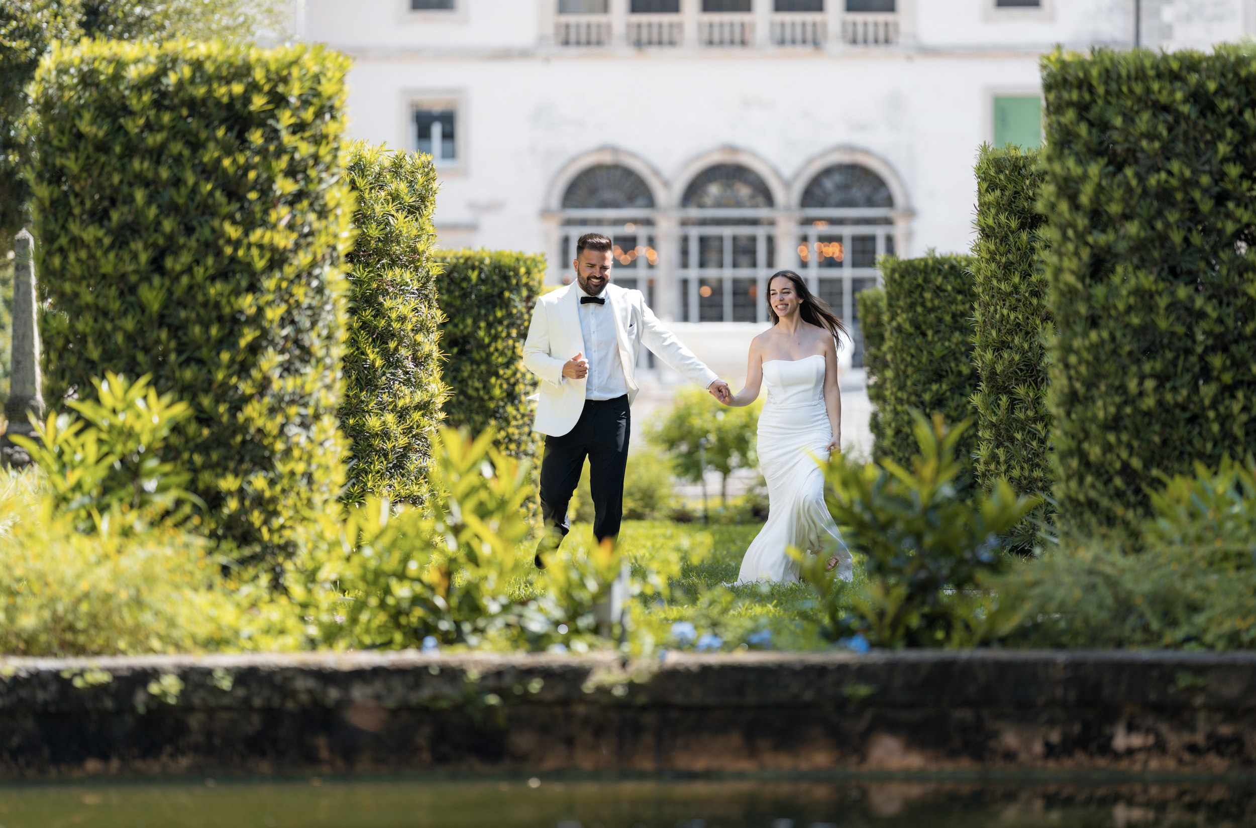A newlywed couple, dressed in wedding attire, walking hand in hand through a lush garden with well-manicured bushes and colorful flowers, smiling and enjoying each other's company.