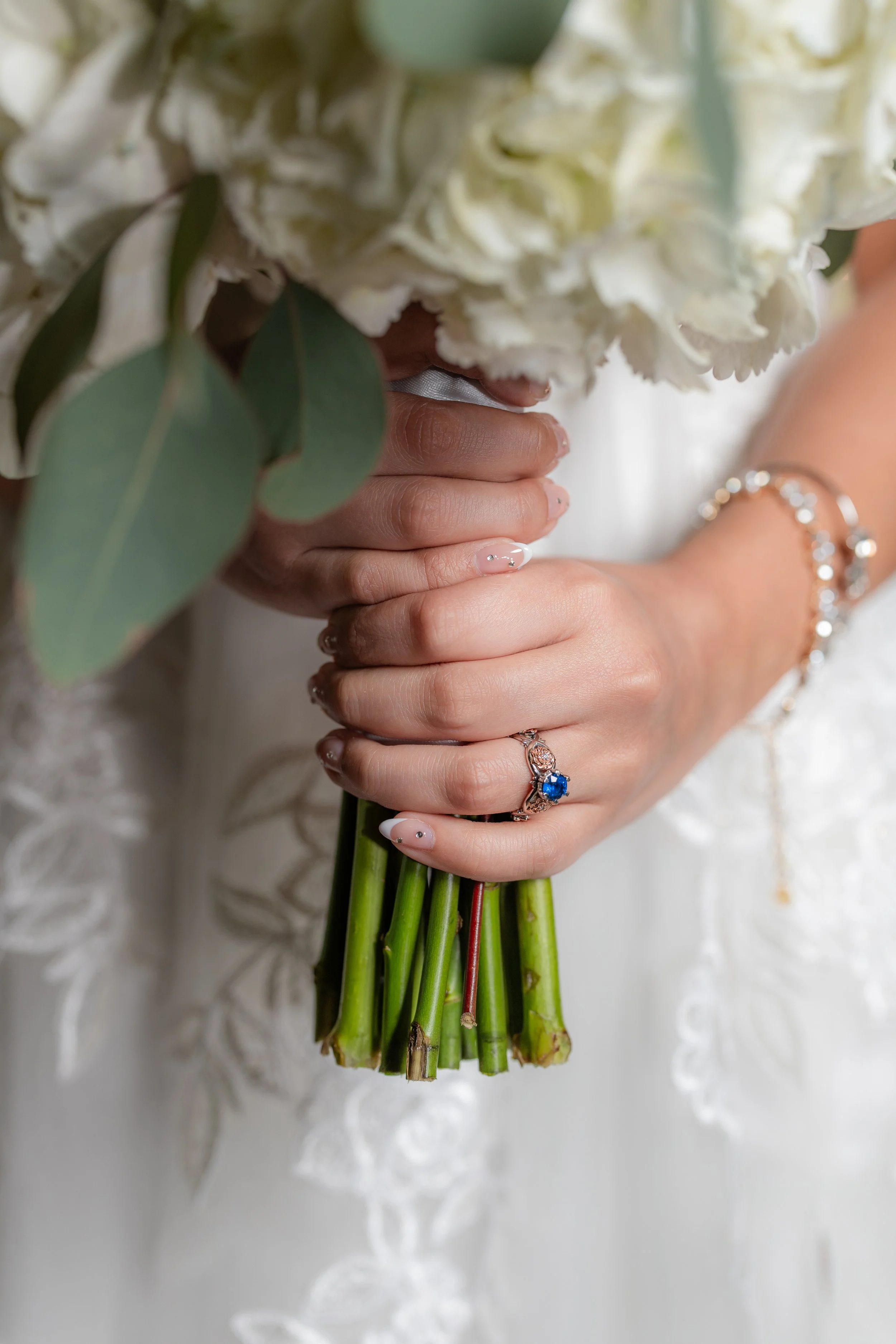A bride holding a bouquet of white flowers with green leaves, showcasing her wedding rings and jewelry.