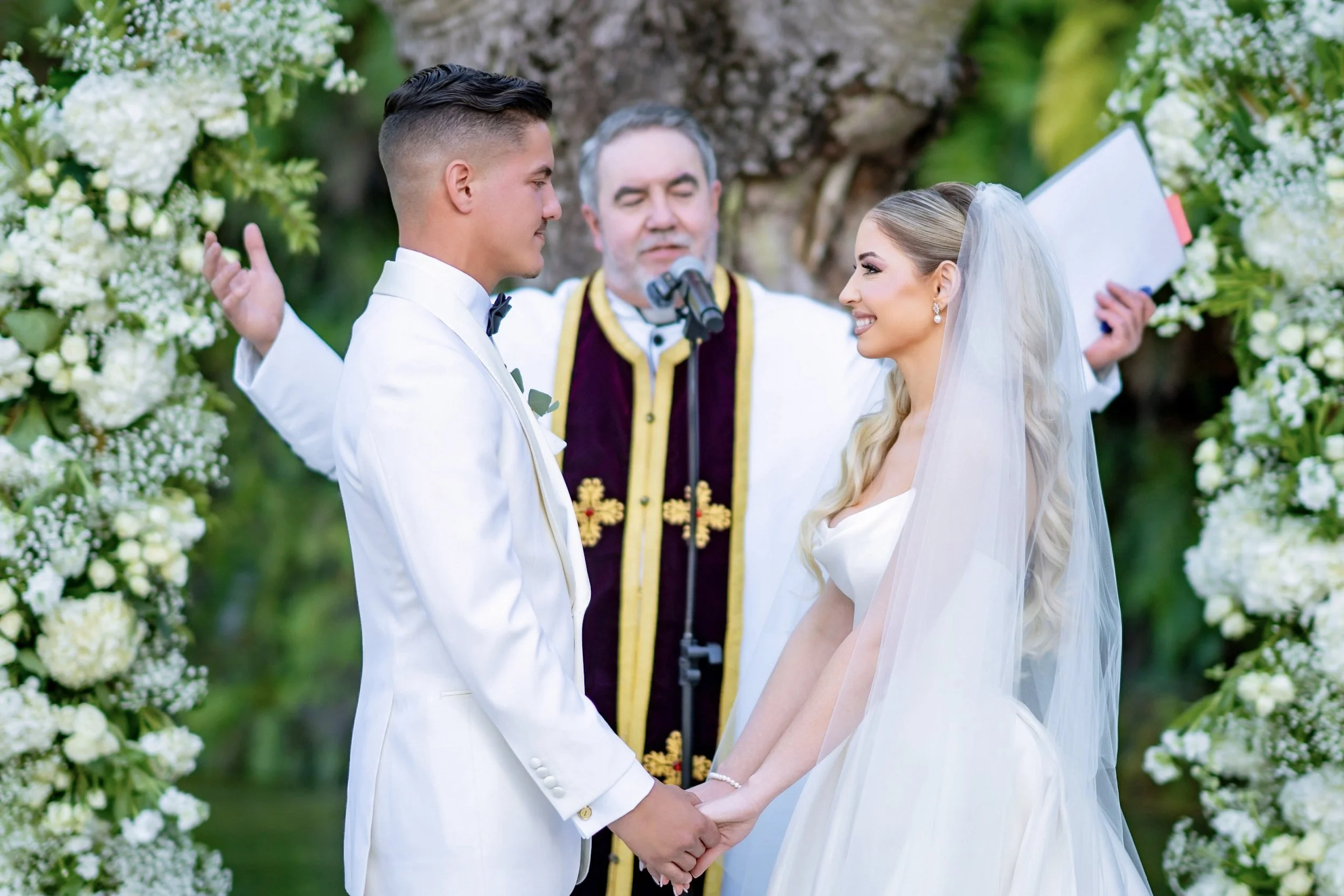 A bride and groom holding hands, facing each other with smiles, during their wedding ceremony outside. An officiant stands behind them, reading from a book, with decorative flowers and greenery in the background.