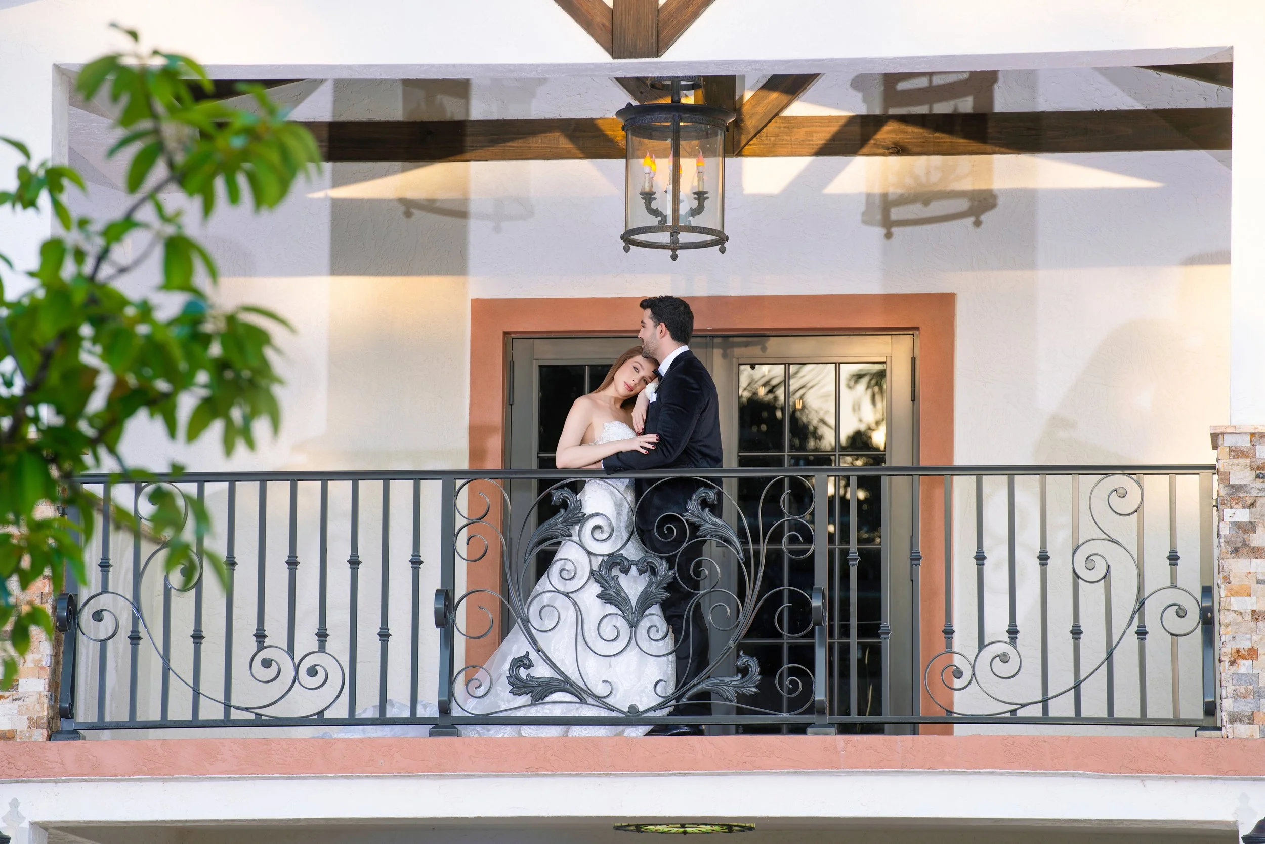A bride and groom sharing a romantic moment on a balcony at sunset with trees reflected in the glass door behind them.