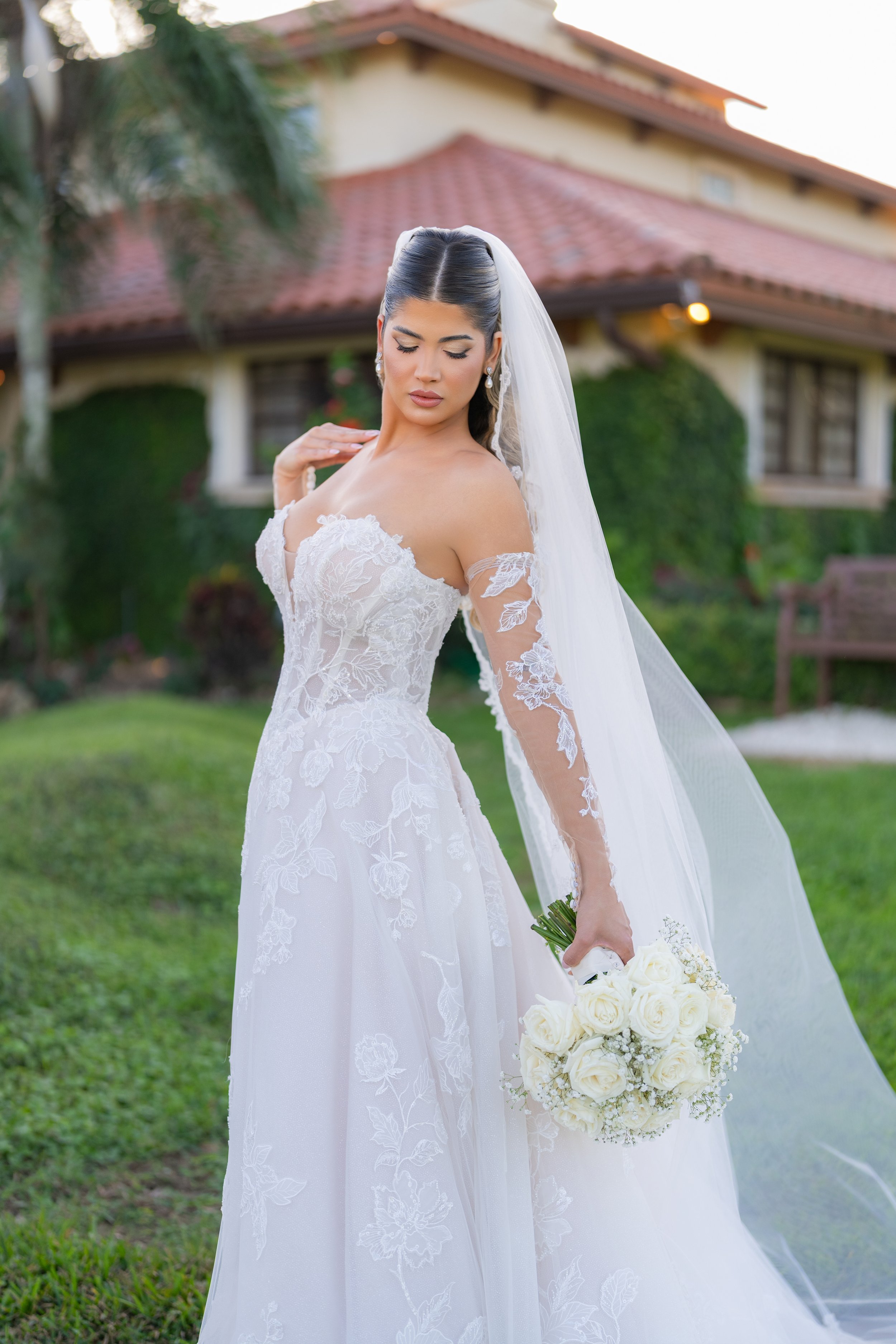 A bride in a white lace wedding gown and veil holding a bouquet of white roses outdoors in front of a house covered with greenery.