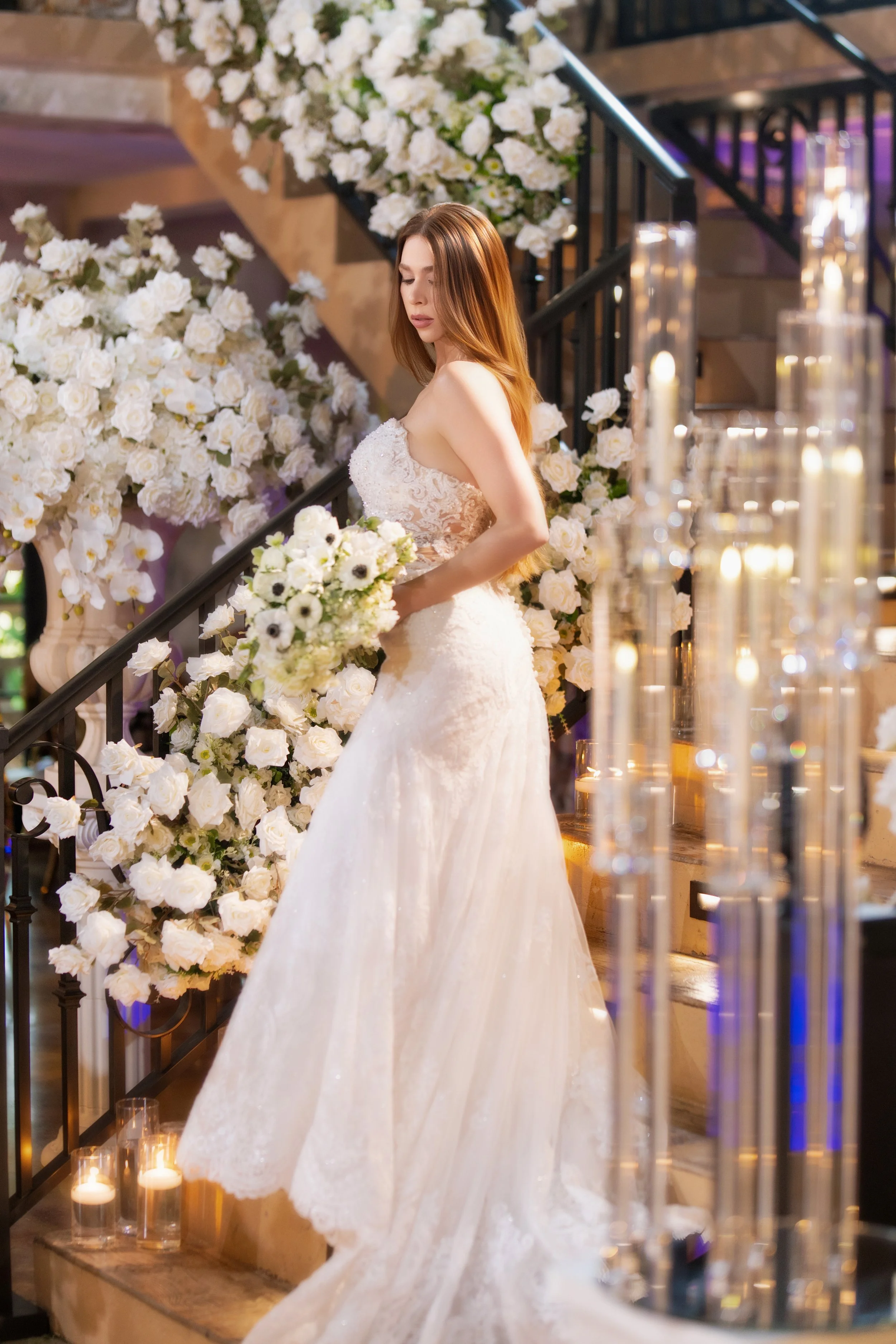 A bride in a white wedding dress holding a bouquet of white flowers, standing on a staircase decorated with abundant white roses and candles in a decorated venue.