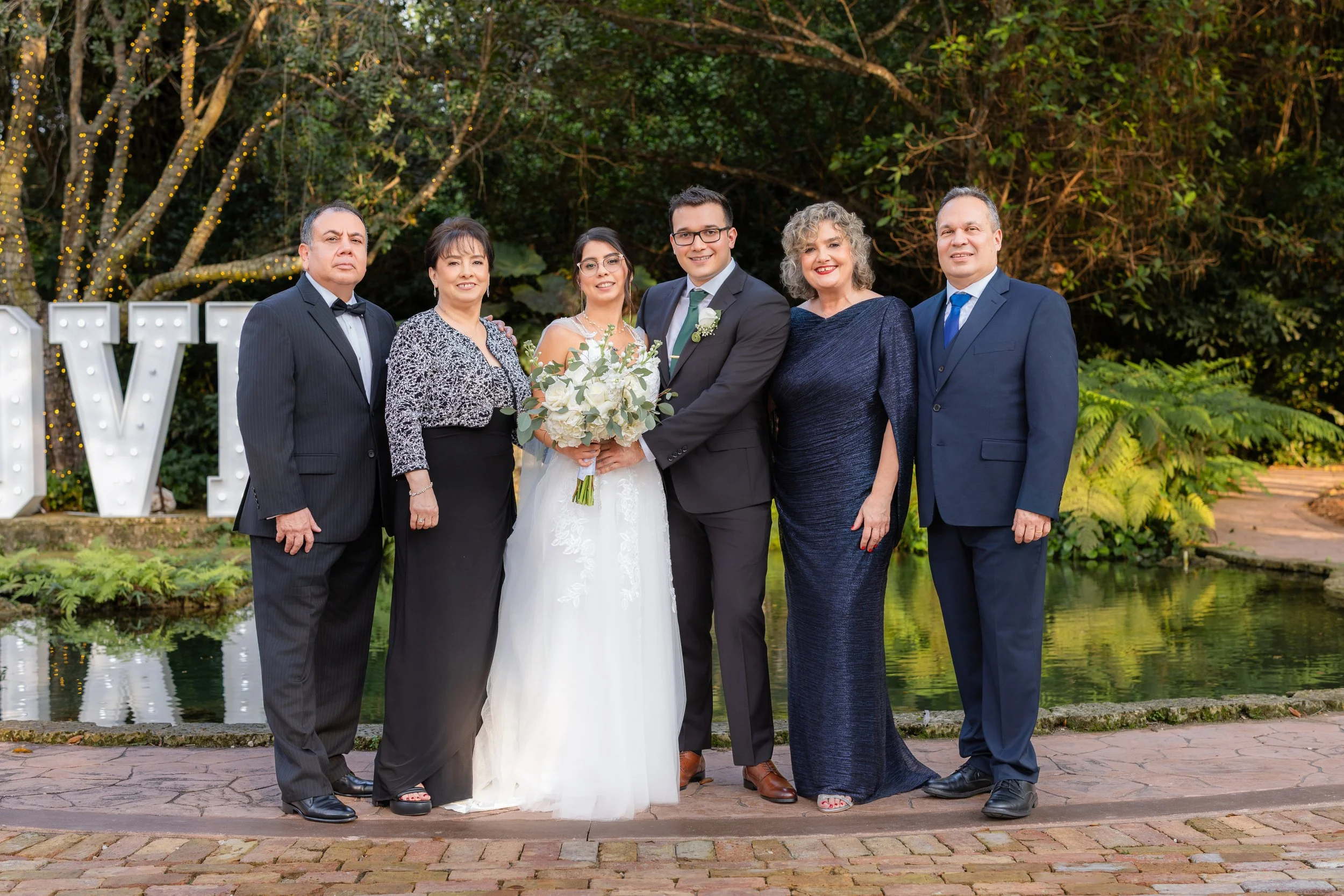 Six people dressed in formal attire standing outdoors near a pond with greenery in the background. A bride in a white wedding dress holding a bouquet, and a groom in a suit in the center. Large illuminated letters spelling "V" and "L" are visible beh