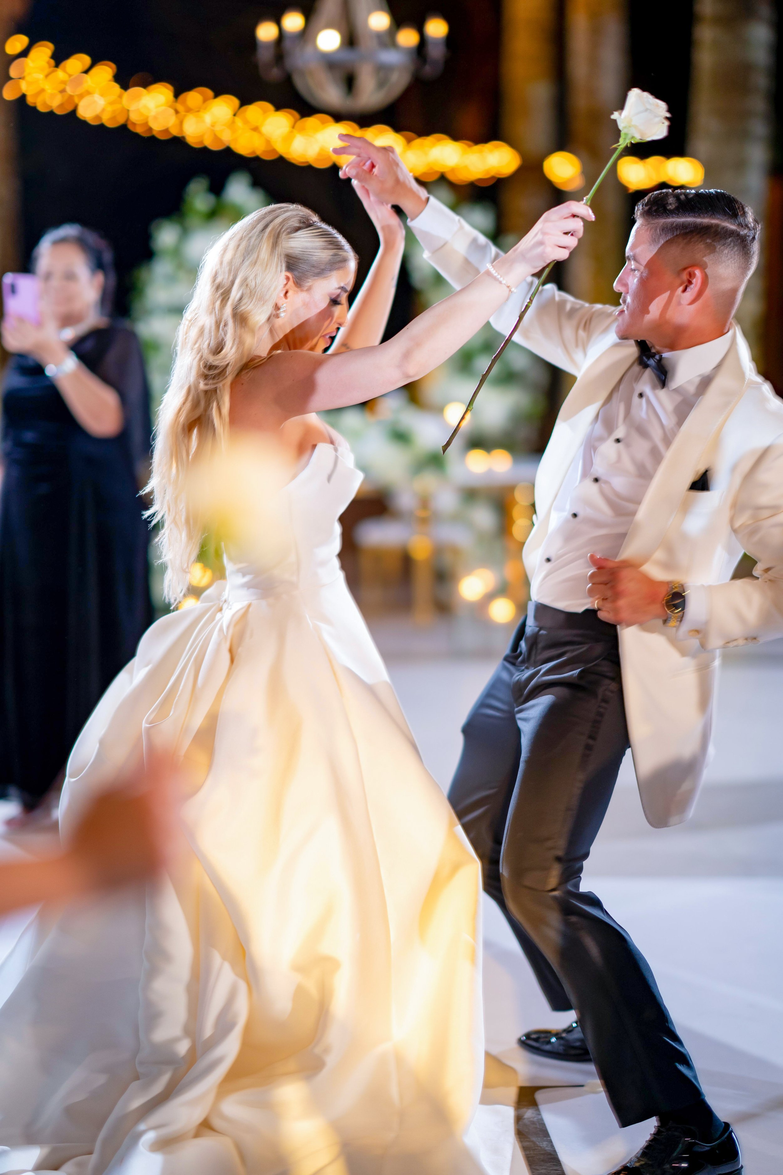 A bride and groom dancing at their wedding reception, with the bride holding a white rose and the groom in a white tuxedo jacket. A woman in black is taking a photo in the background.