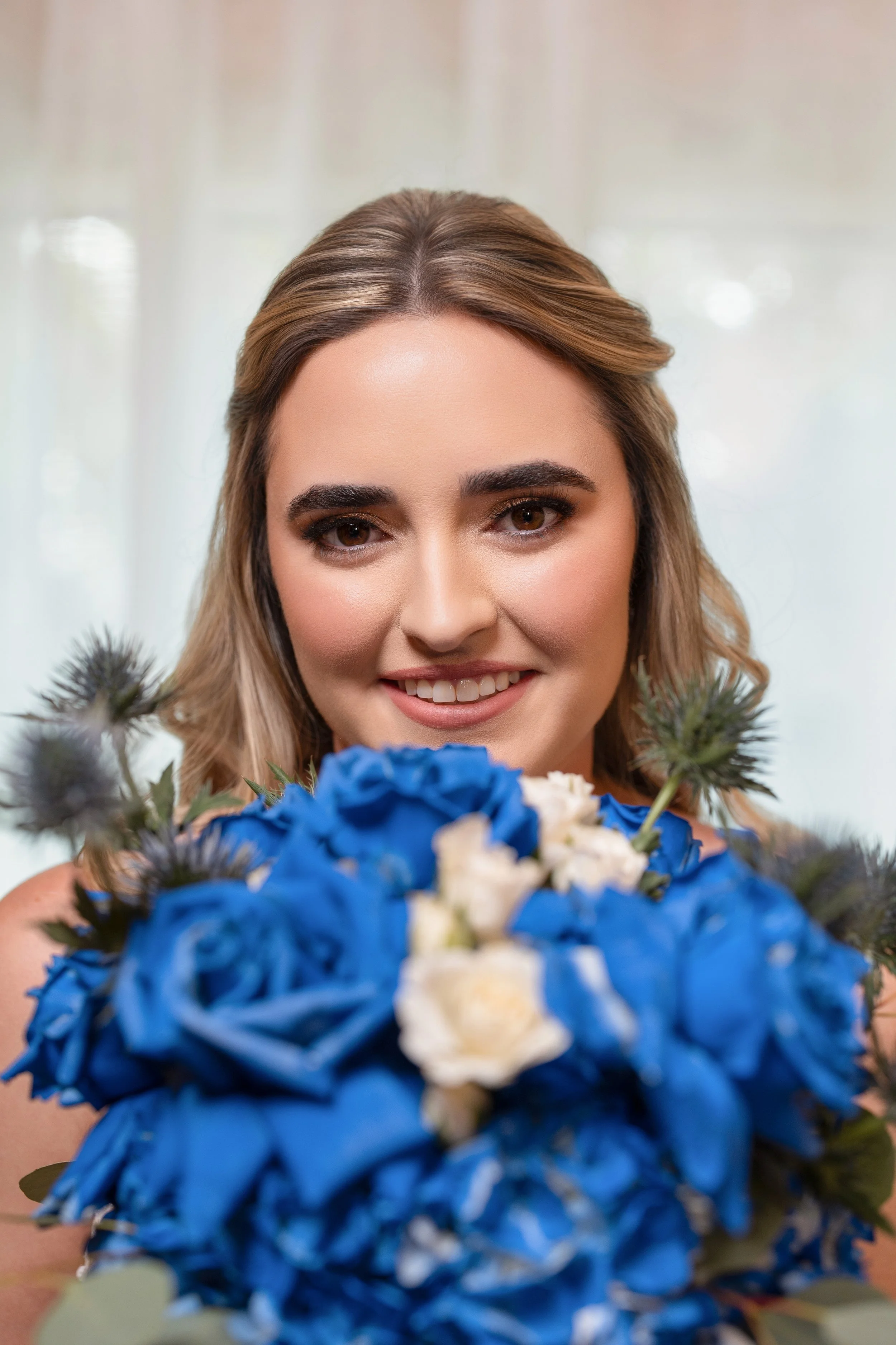 A woman with light brown hair and makeup smiling while holding a bouquet of blue and white flowers.