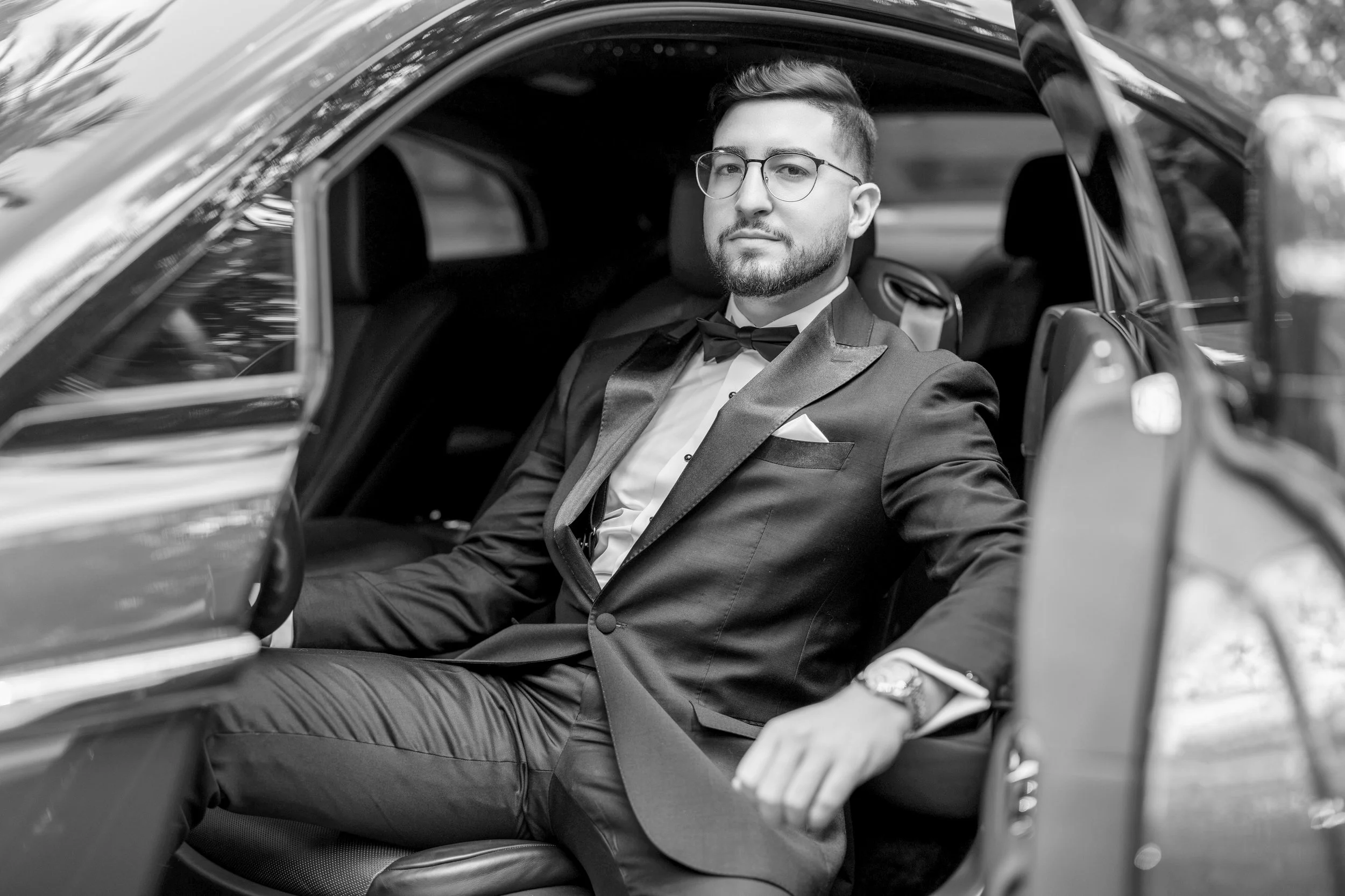 A man in a tuxedo sitting in the back seat of a car, looking at the camera with a confident expression.